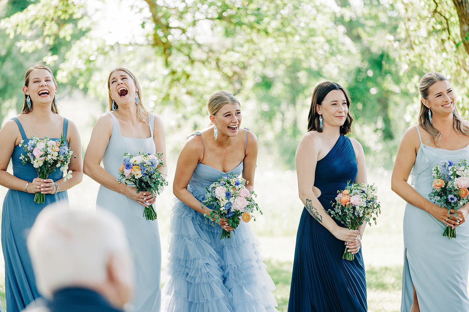 Five bridesmaids in various shades of blue dresses stand outdoors, holding colorful bouquets and laughing joyfully during a wedding ceremony, beautifully captured by an Eau Claire Wedding Photographer amid lush trees and greenery.



