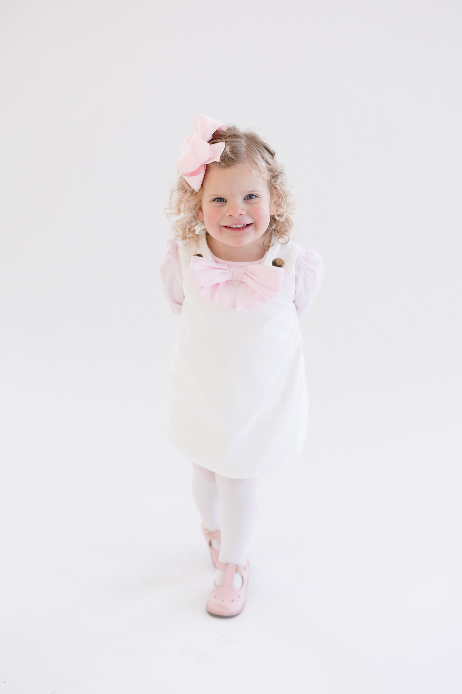 Little girl wearing a white and pink dress with her hands behind her back as she smiles at the camera
