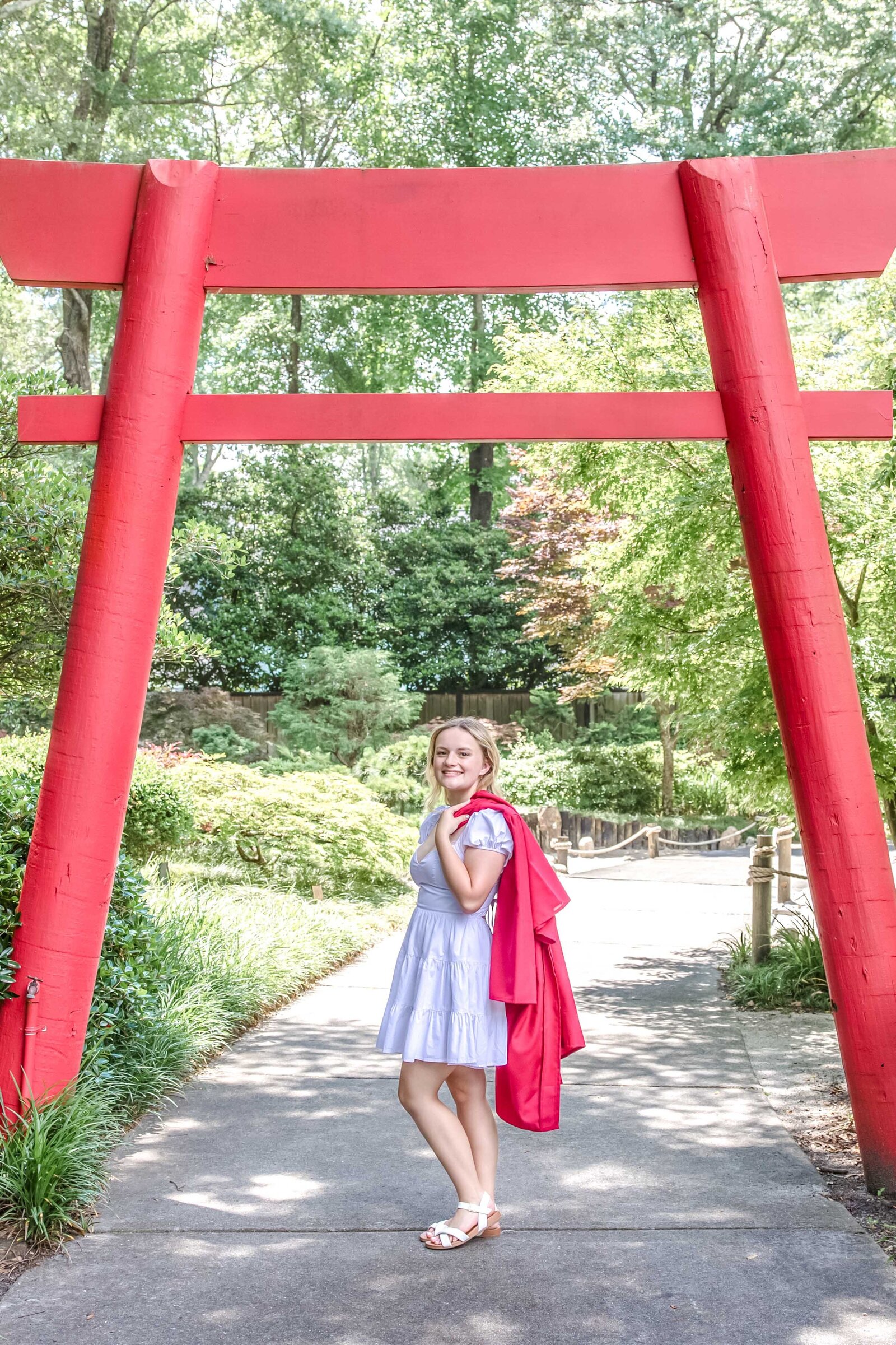 A high school senior holding her graduation gown over her shoulder, posing under a vibrant red Torii gate at the Arbortum in Wilmington, North Carolina. This unique setting highlights the charm of destination graduation photography, perfect for capturing distinctive senior photos.