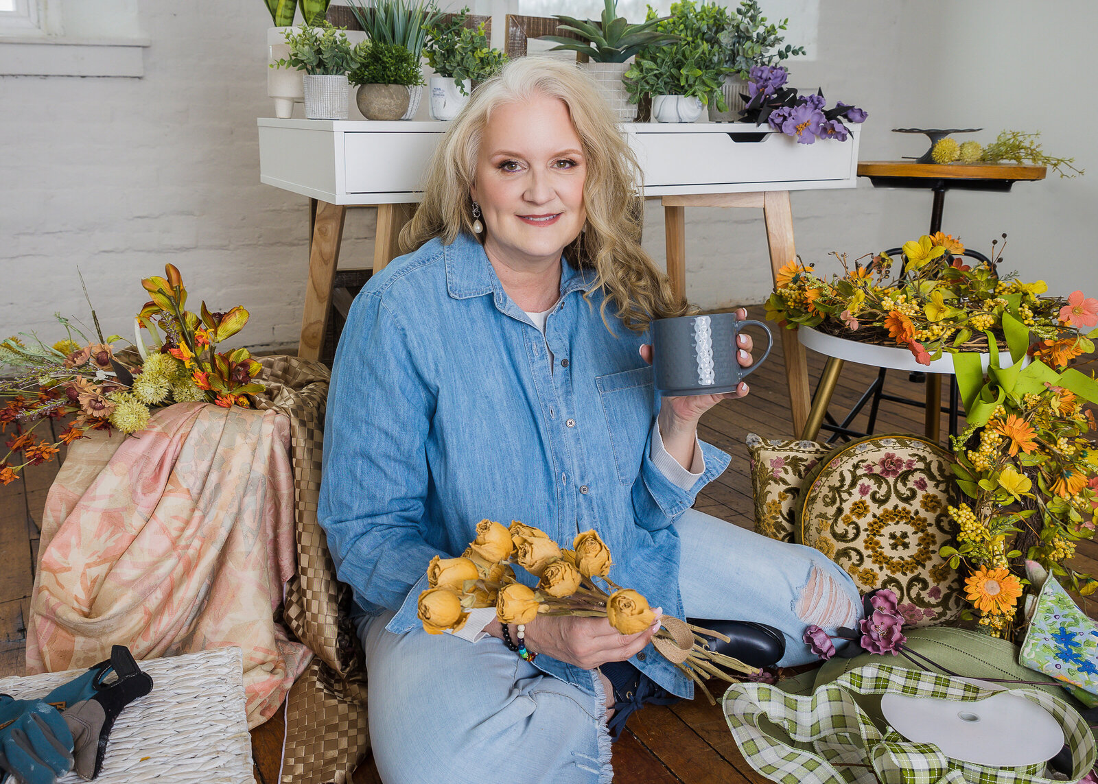 woman sitting surrounded by fake flowers and crafts holding a cup of coffee