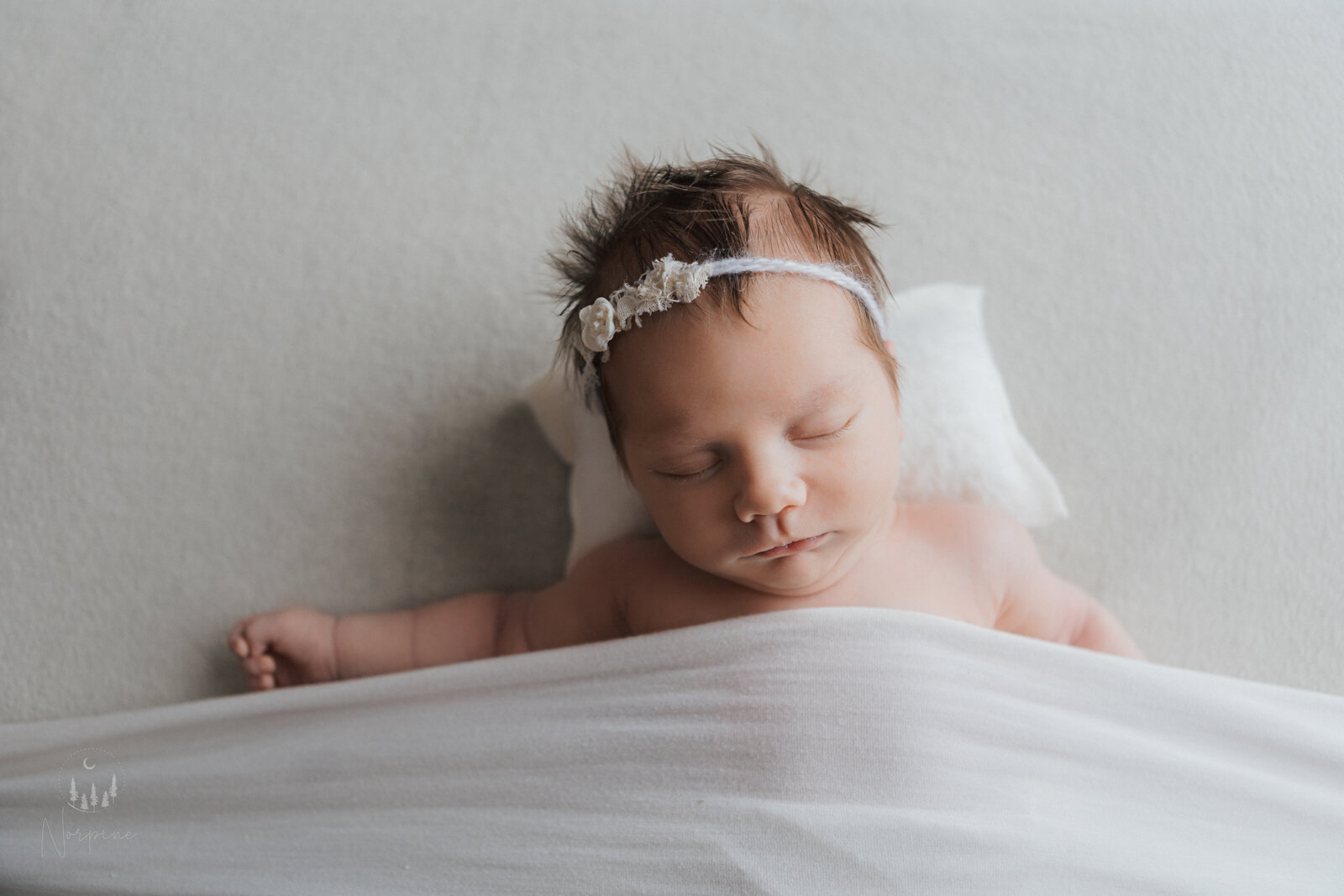 a newborn girl in the tucked in pose with a white background, blanket, and pillow, wearing a white headband. She is relaxed in posture with eyes closed.