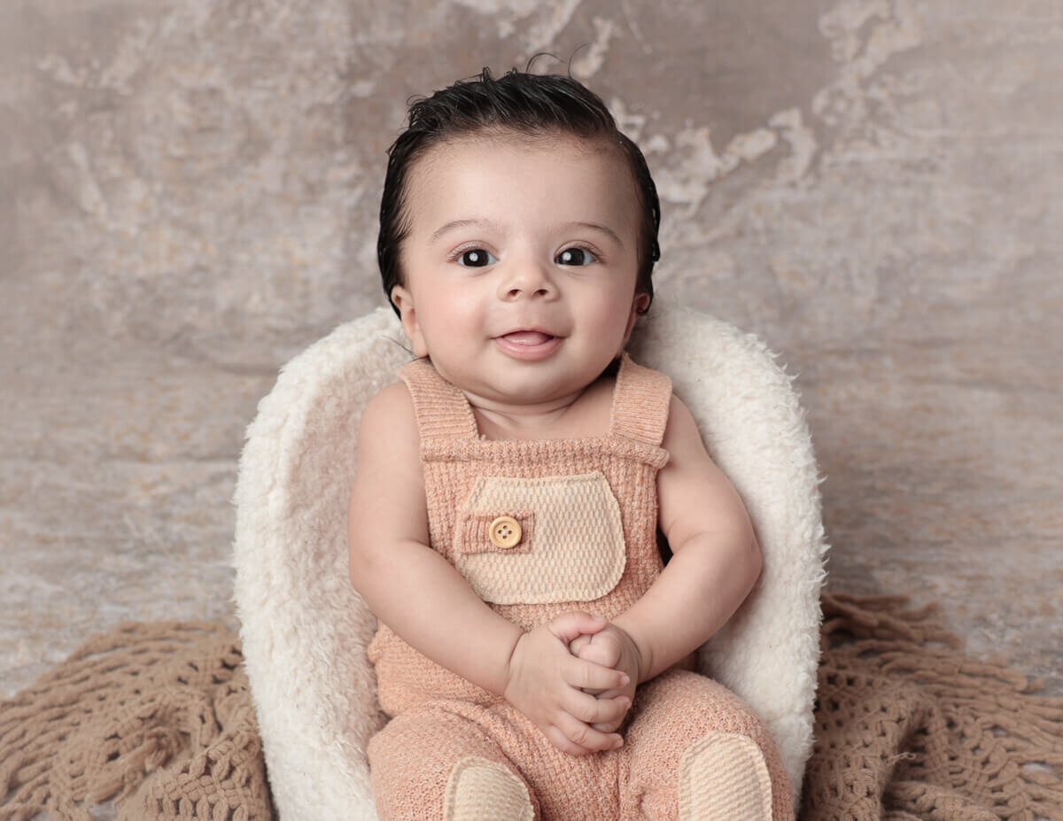 A three-month-old baby sitting comfortably in a white soft chair during a milestone photography session in a studio in Rochester, NY. The baby is smiling, showcasing their adorable features and capturing the joy of early childhood milestones.