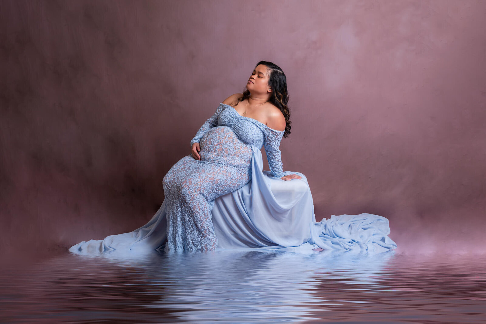 A pregnant woman reclines elegantly on a white bench, wearing an off-the-shoulder light blue lace gown, radiating peace and anticipation.