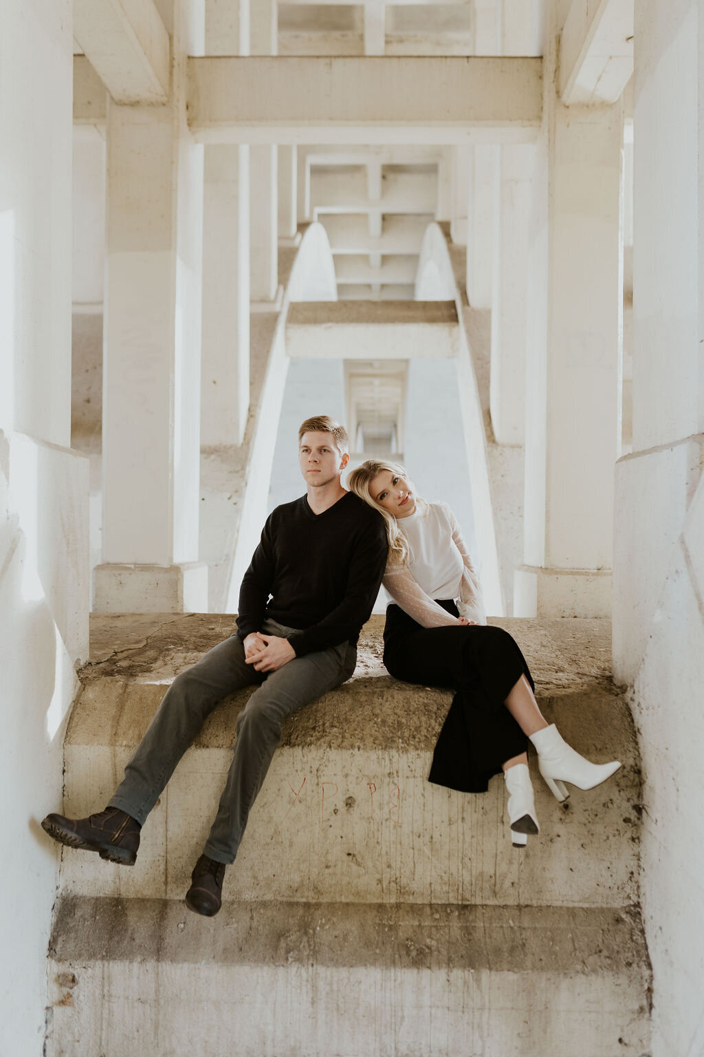 Woman wearing a black skirt and white boots leans on man with a black sweater while they sit on a ledge under a white bridge
