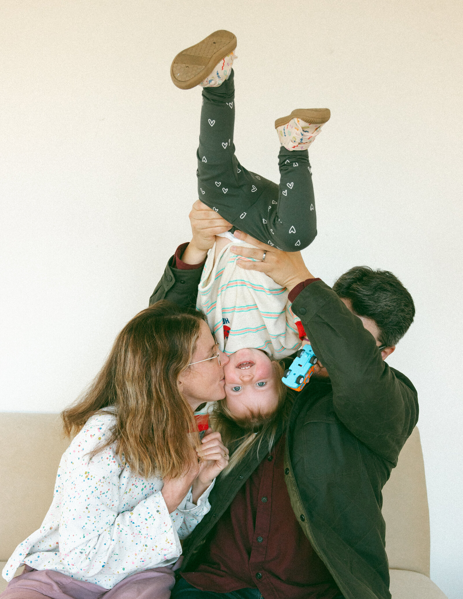 baby being held  upside down in the air by dad while mom kisses her cheek