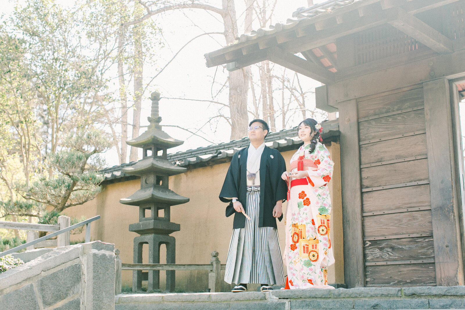 Bride in floral wedding kimono rental and groom in black montsuki walking on a wooden bridge surrounded by spring blossoms, The Huntington Library Japanese Garden, Los Angeles California | 花嫁が花柄打掛レンタル、新郎が黒紋付で春の花に囲まれた橋を歩く