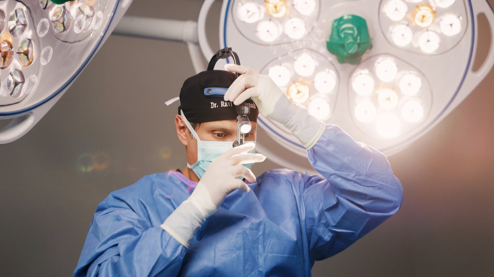 Surgeon in a blue gown adjusting his headlight under operating room lights