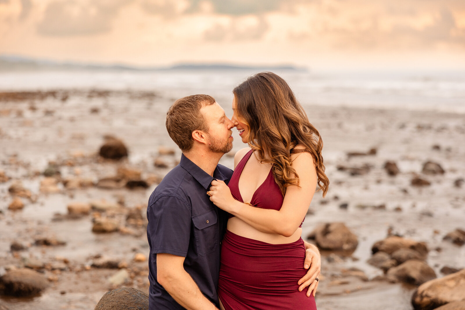 woman in red sits on man's lap on rugged rocky beach coastline