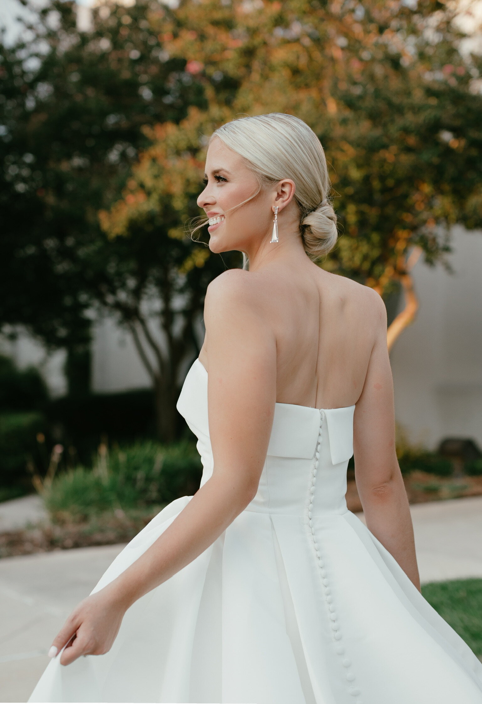 Woman in a strapless white gown, embodying natural glam wedding makeup and effortless wedding hairstyles, smiling and turning gracefully with trees in the background.