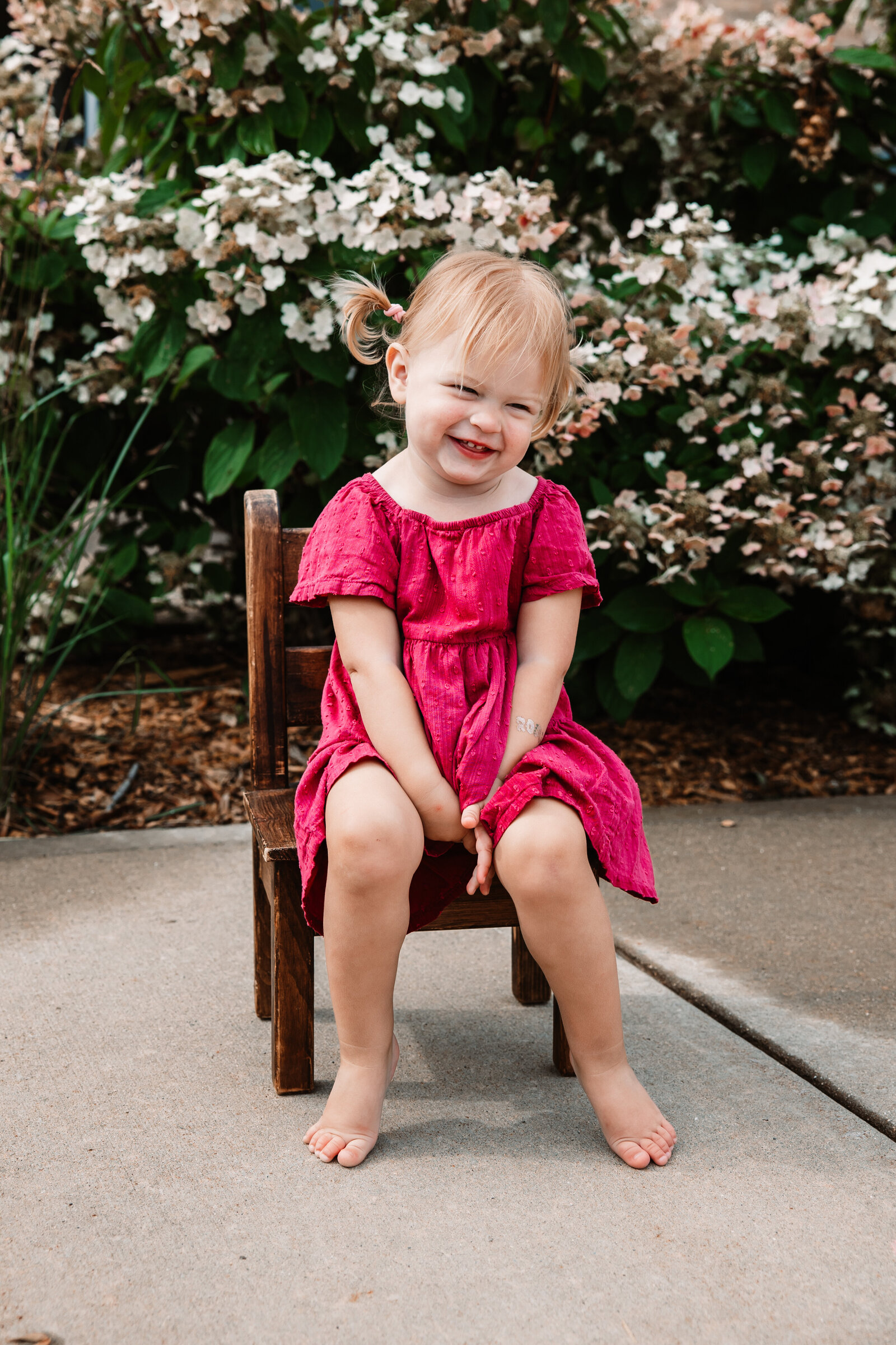 A toddler girl in a pink dress sits in a wooden chair in a garden sidewalk giggling
