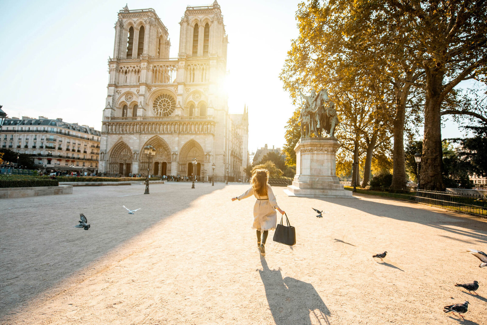 A person with long hair runs towards Notre-Dame Cathedral in Paris, holding a large black bag. The sun shines brightly, casting a warm glow. Pigeons fly across the plaza, and a statue stands nearby surrounded by trees—an ideal scene for a European travel specialist to capture in their itinerary planning.