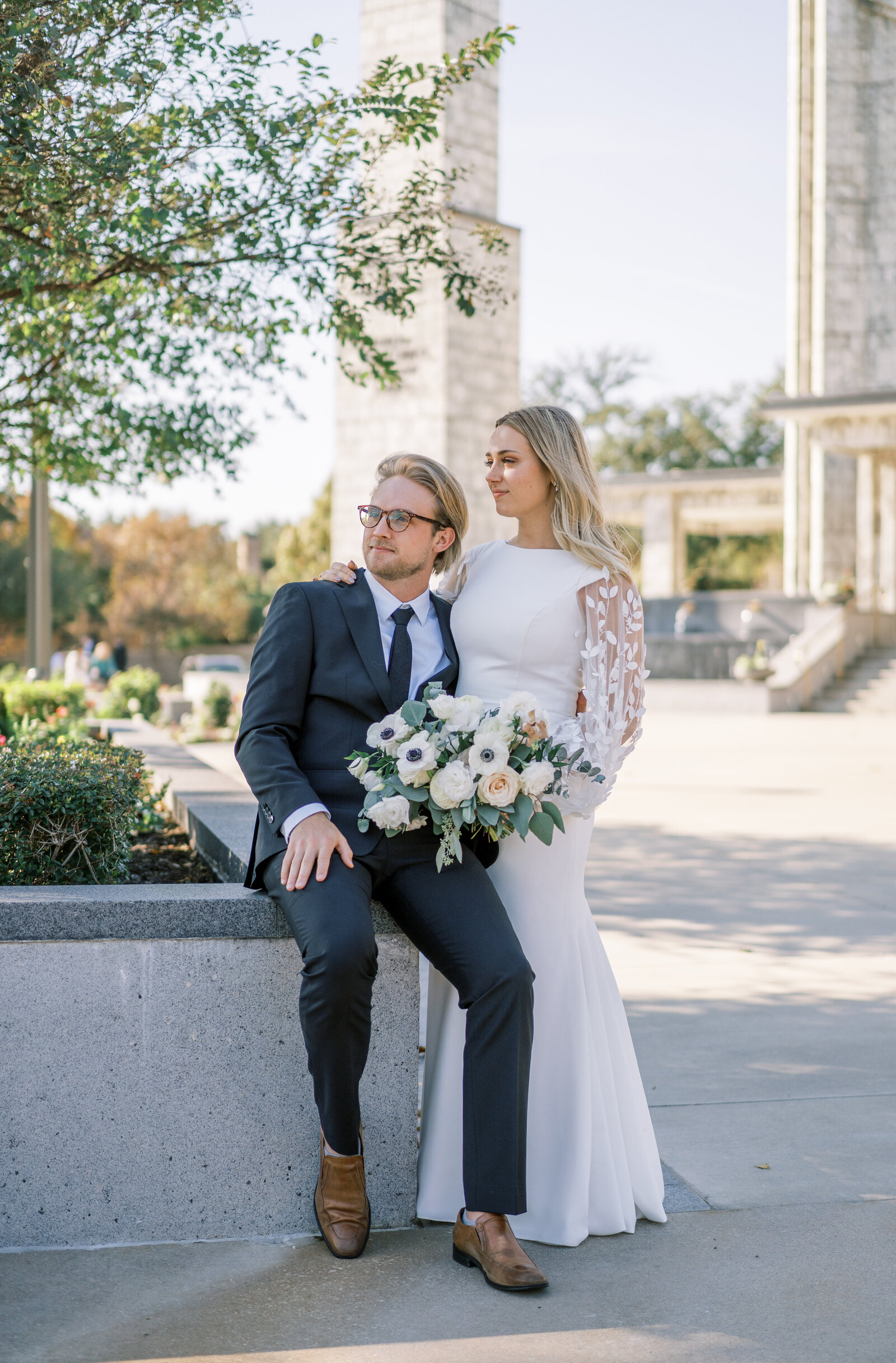 Portrait of bride and groom in a white wedding gown and black suit sitting by a garden in a courtyard.