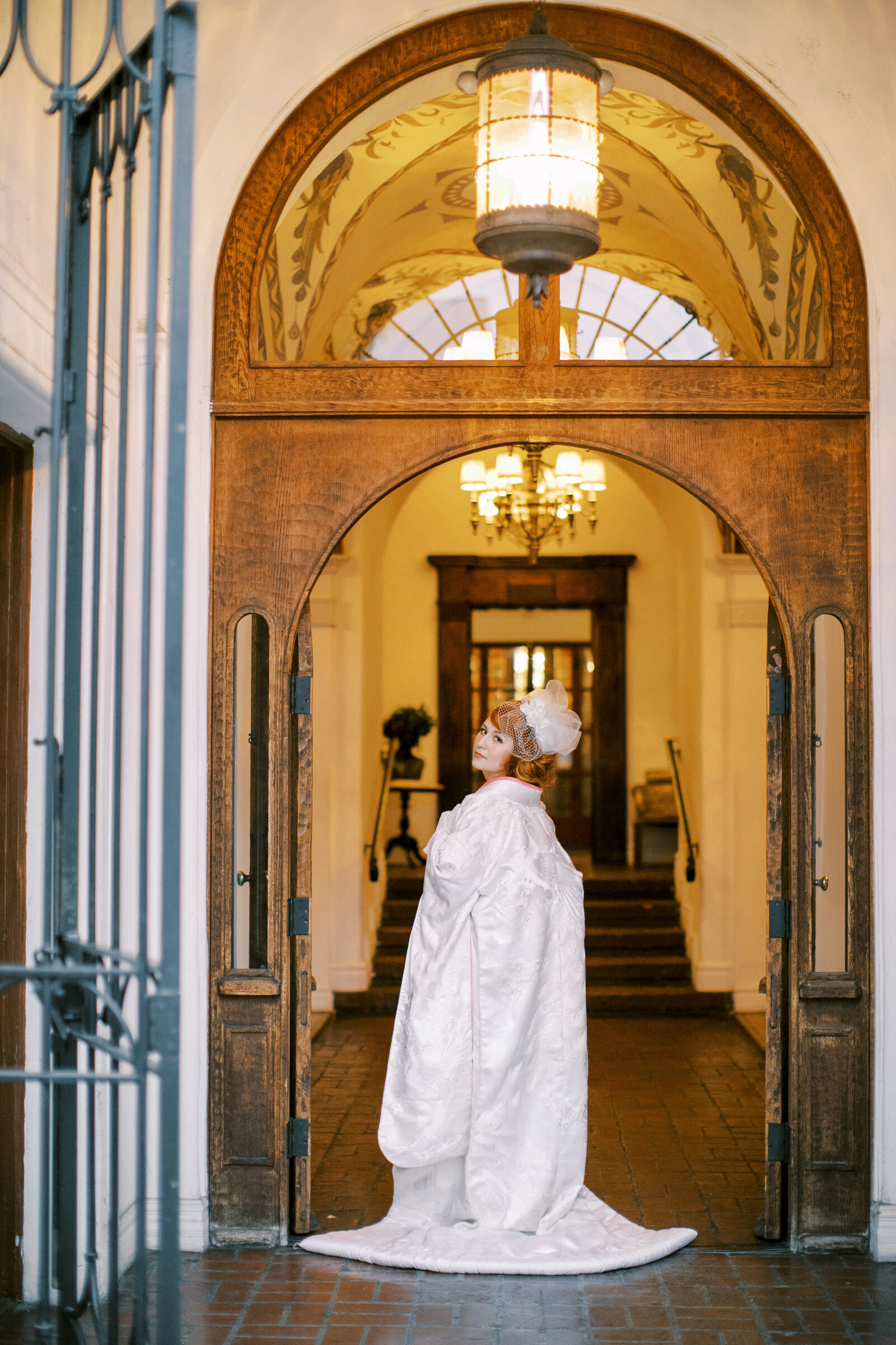 Bride standing gracefully under the arched doorway in a white shiromuku with soft pink accents at Carondelet House, Los Angeles California. Available for wedding kimono rental in Los Angeles | ロサンゼルス・カロンデレットハウスのアーチ下で佇む、淡いピンク掛下の白無垢花嫁。着物レンタル可能。