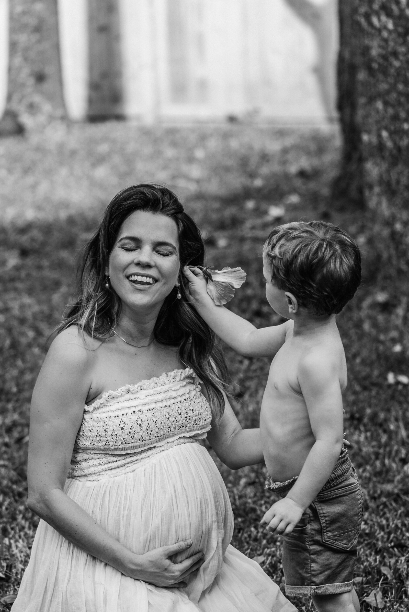 black and white photo of a pregnant mother laughing while her  her toddler boy (without shirt) puts a flower behind her ears.