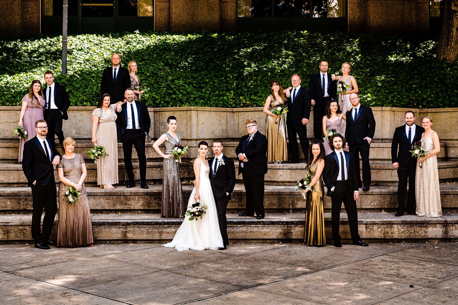 large wedding party in dark suits and metallic dresses poses in downtown Durham, North Carolina
