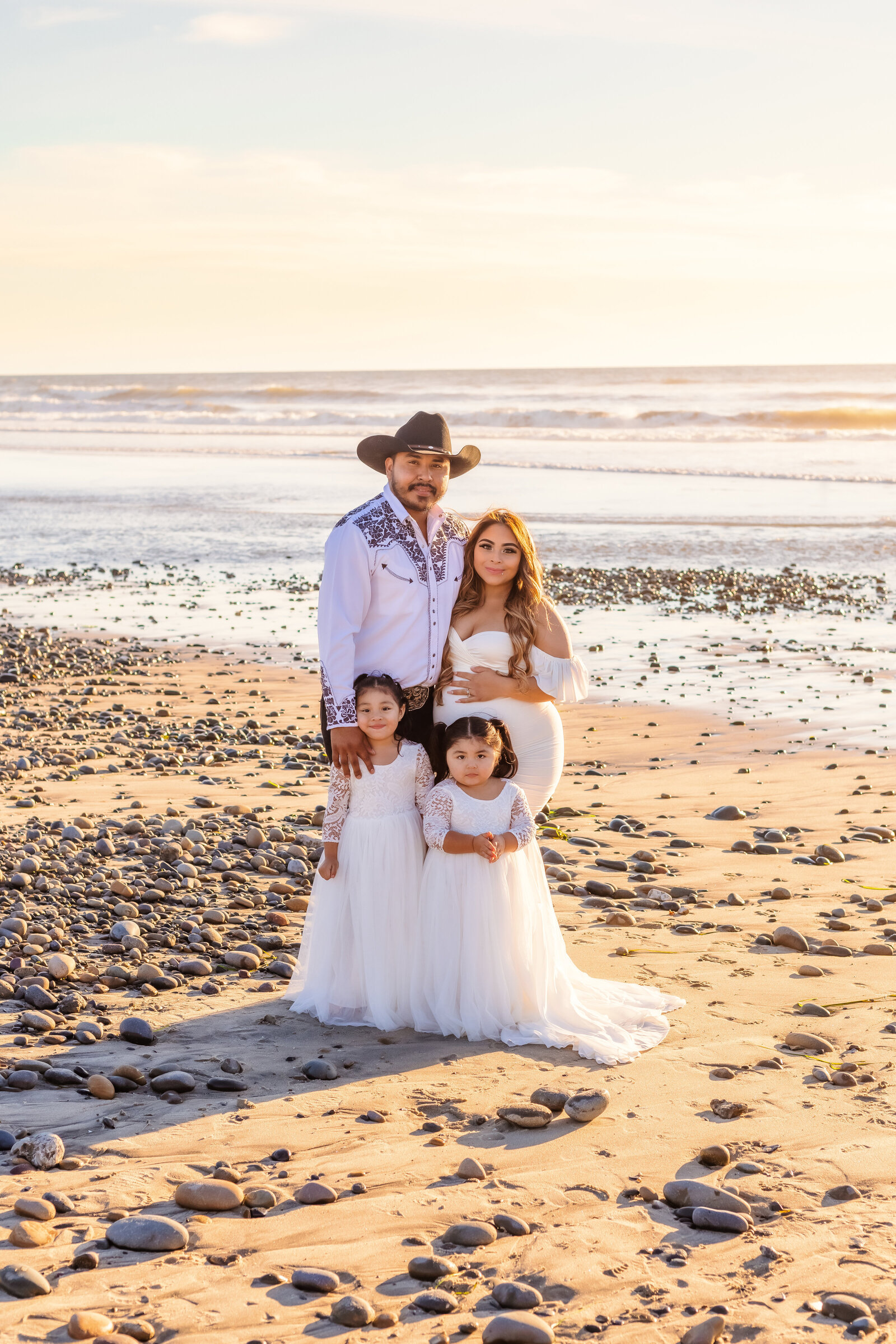Maternity Photographer, a young family stands together at the beach, dad wears a cowboy hat and mom is expecting another child