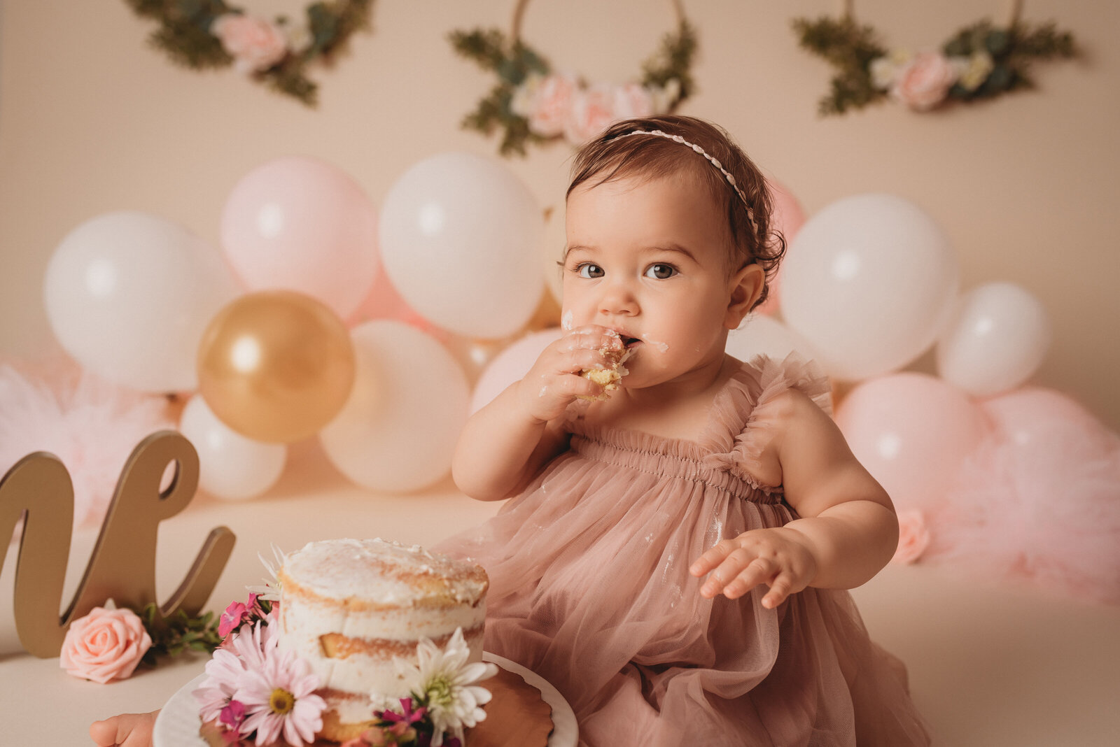 One year old baby girl wearing pink tulle dress sitting on tan backdrop at Atlanta milestone photography studio eating cake and looking at camera with balloons and flowers behind her