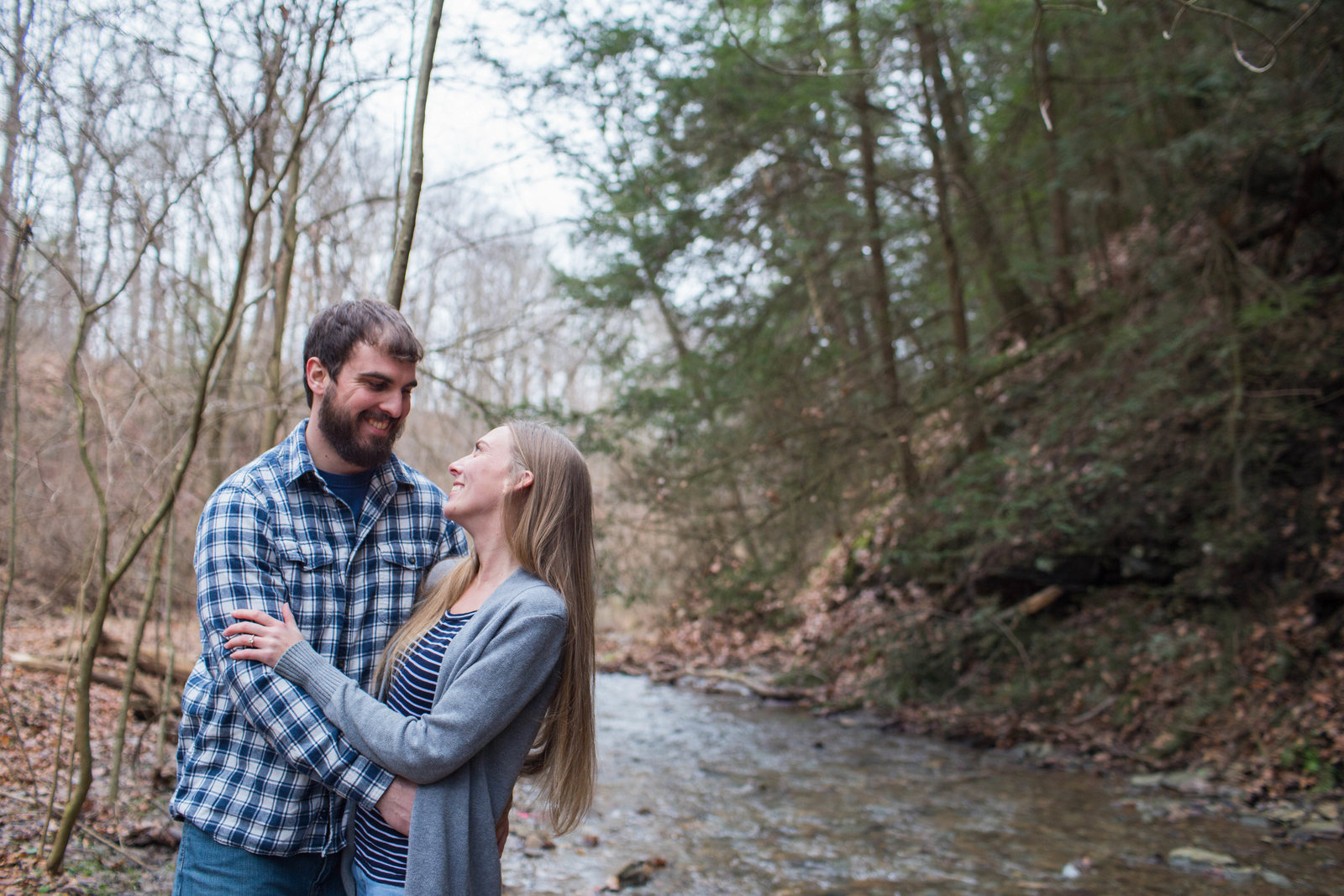 engaged couple hugs beside a creek running through hartwood acres