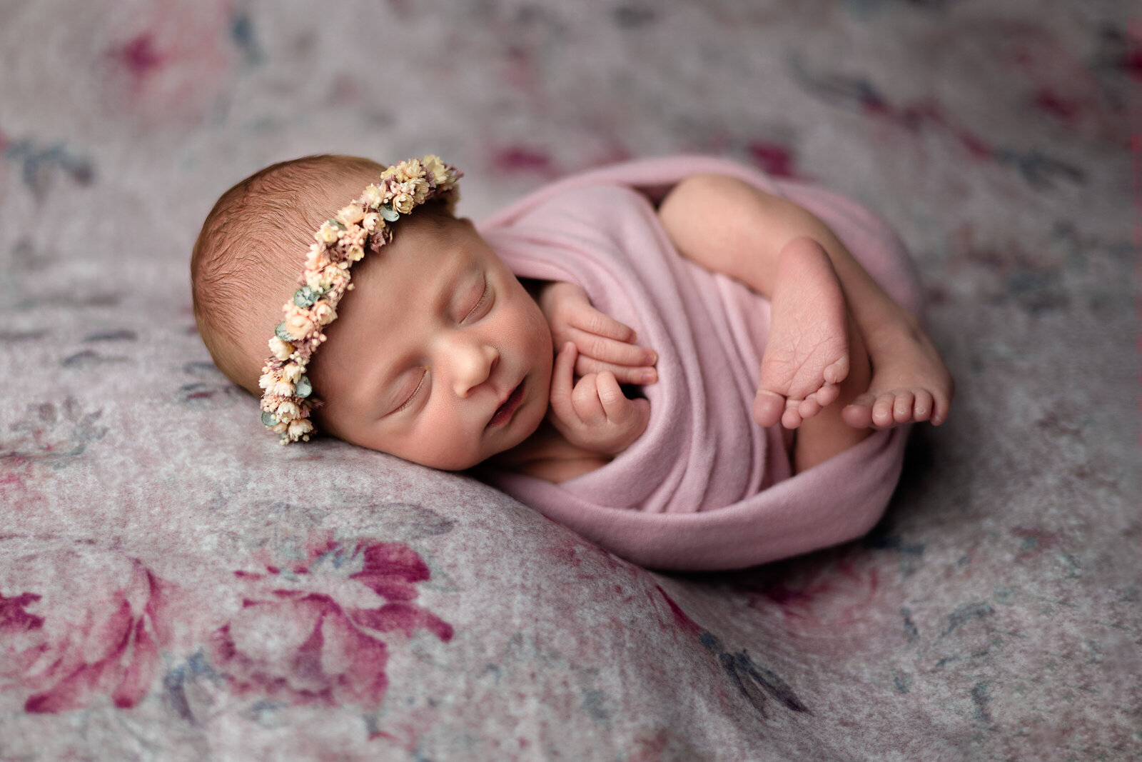 Newborn girl swaddled in pink with a matching floral halo, posed on a gray and pink floral background