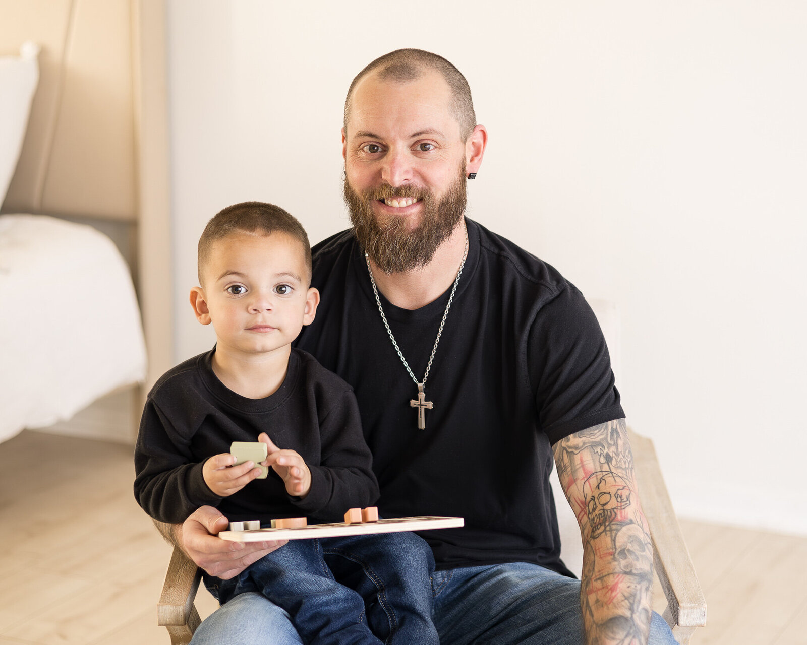 A father and son sitting in a chair in a studio room.  Dad has dark brown hair and beard, wearing a black shirt, blue jeans, and a cross necklace, smiling at the camera with son on his lap.  Son has dark brown hair wearing a black shirt, blue jeans, and looking at the camera 
