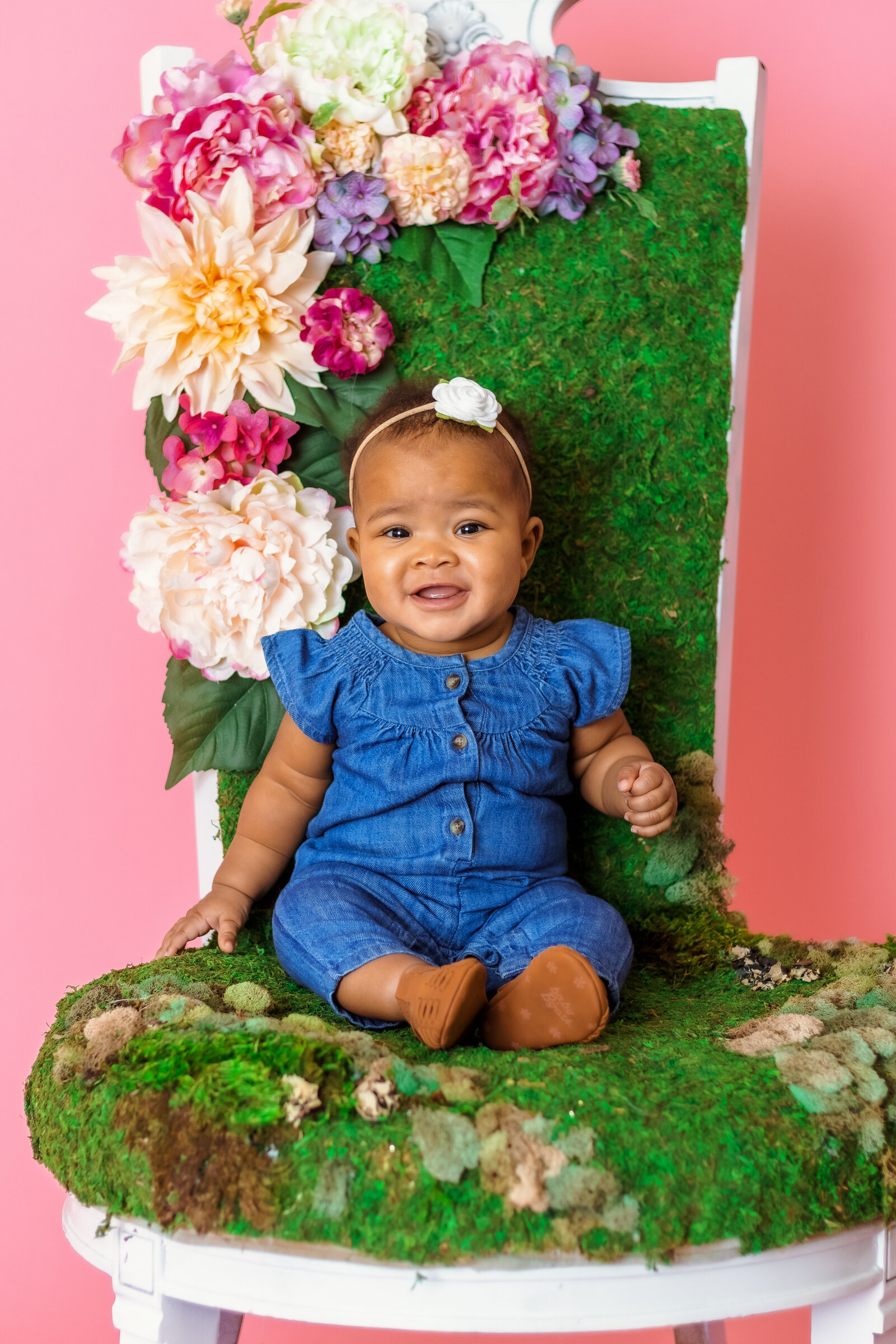 Milestone Photographer, a baby girl sits on a chair with faux flowers and moss covering it