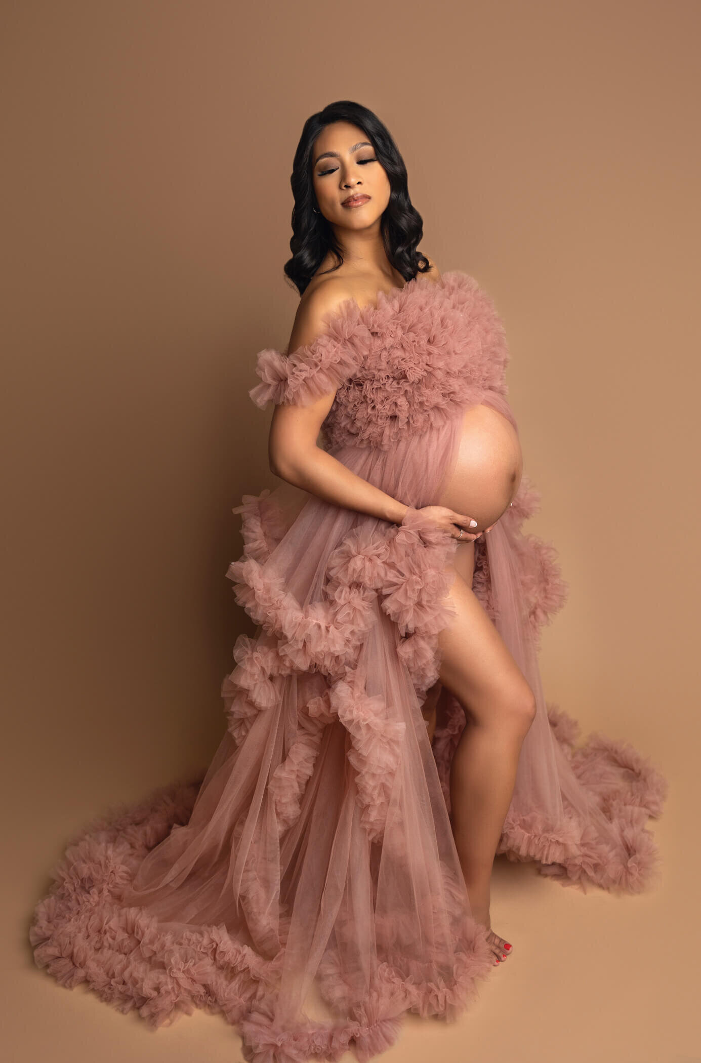 A pregnant woman poses gracefully against a warm beige background, wearing a flowing, off-the-shoulder pink tulle gown with ruffled details. The gown elegantly drapes around her, accentuating her baby bump. She has long, wavy dark hair and a serene, confident expression. Her pose is poised and elegant, exuding a sense of beauty and anticipation.