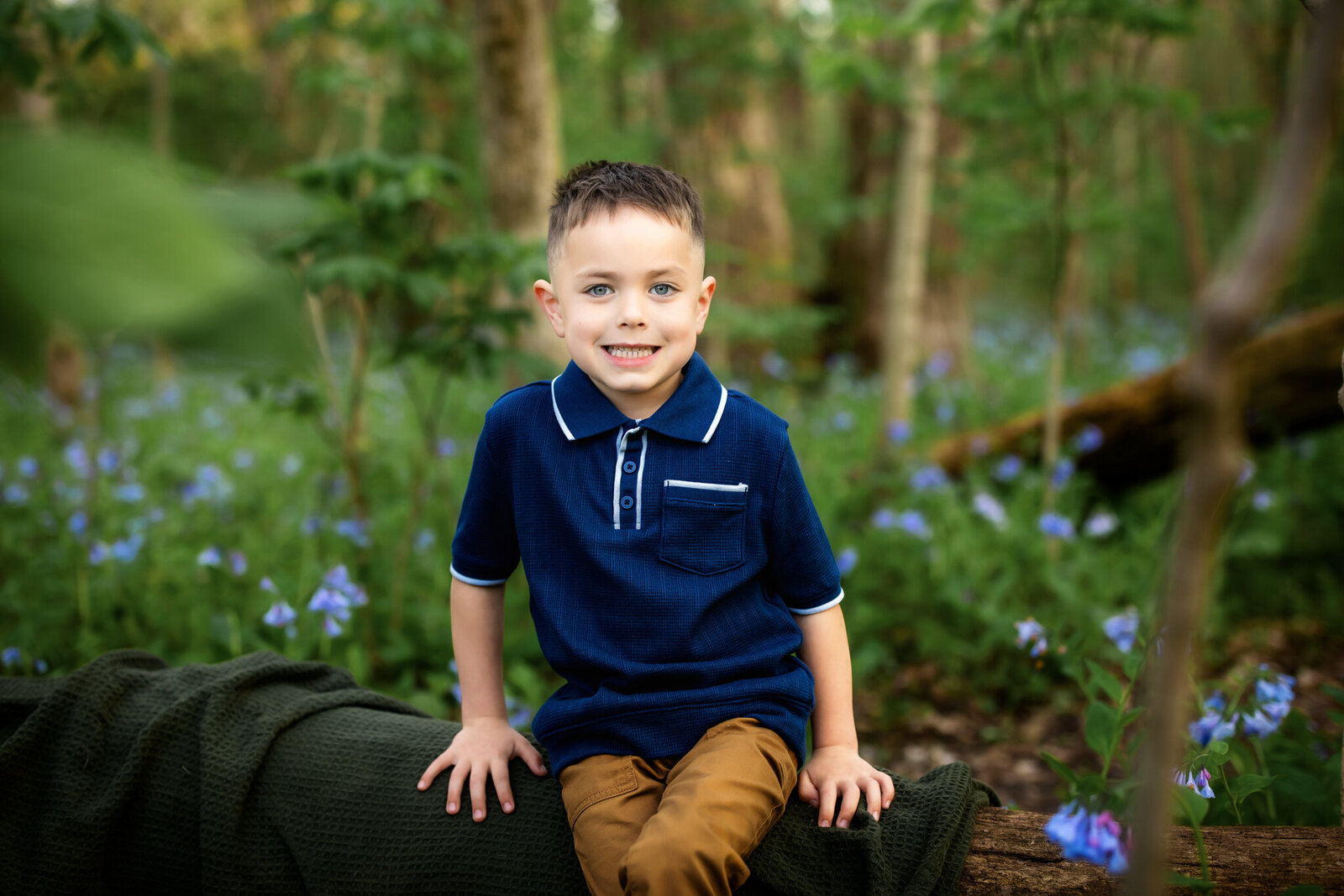 A young boy with short brown hair and blue eyes, wearing a navy blue polo shirt and brown pants, sits on a log in a forest with green foliage and purple wildflowers, smiling at the camera during outdoor portraits by a Bloomington family photographer.
