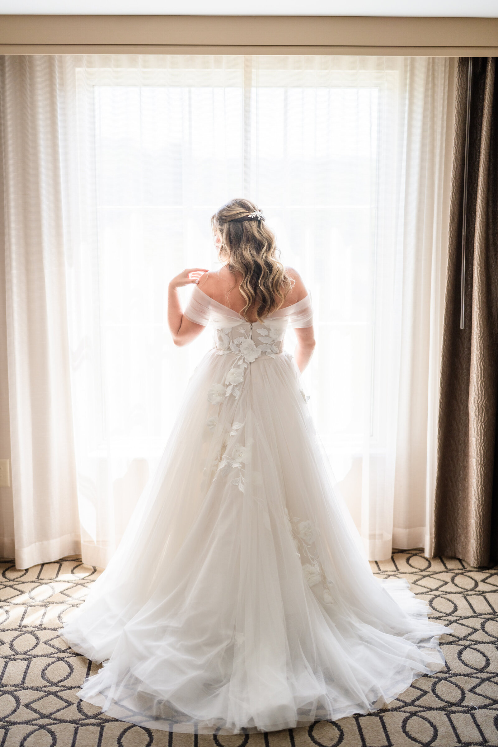 Bride shows off the details of her wedding dress while standing in the window of her suite at the Hilton