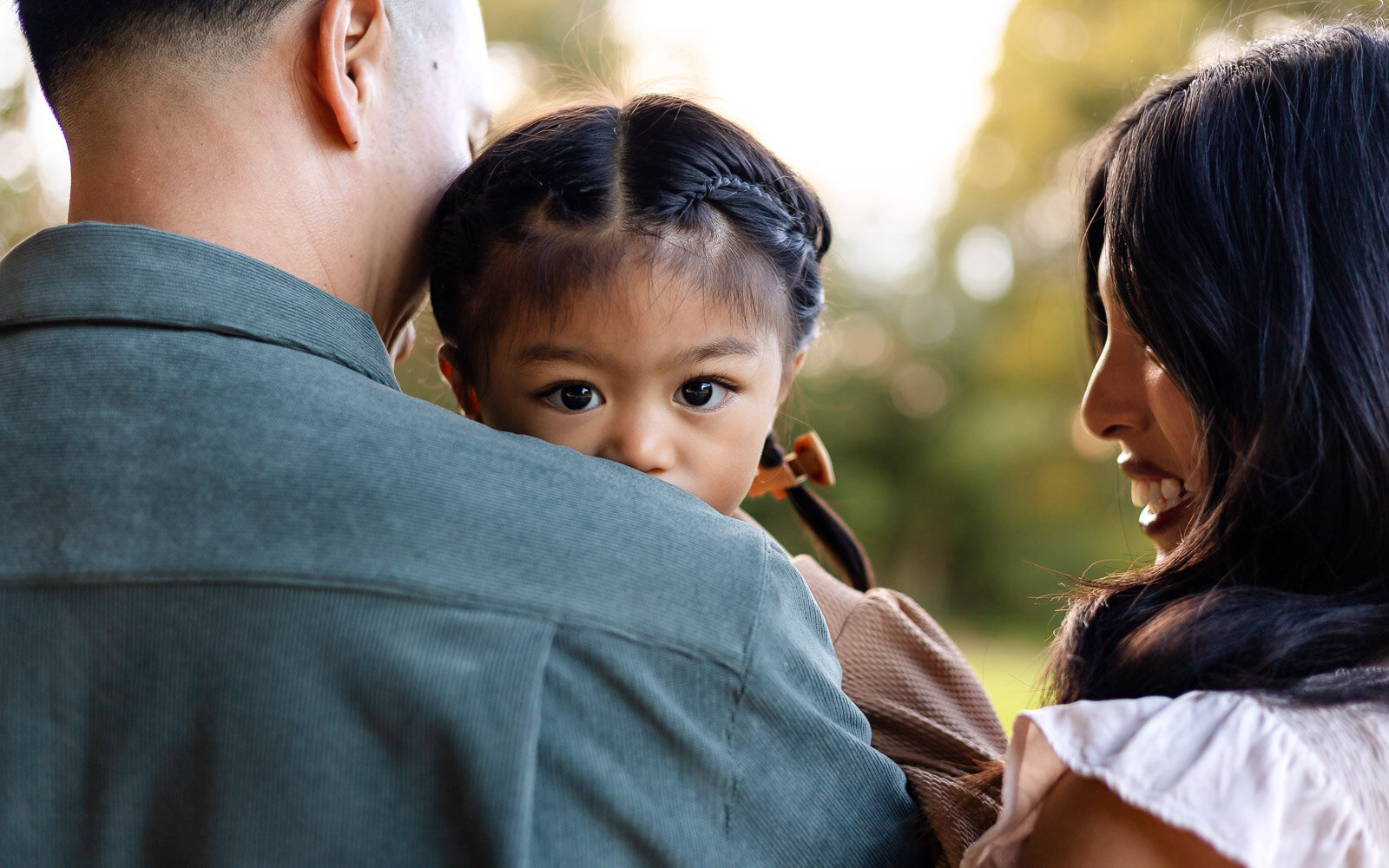 Toddler girl looking over dad’s shoulder during a family moment  by Ellobelle Photography Family Photographer