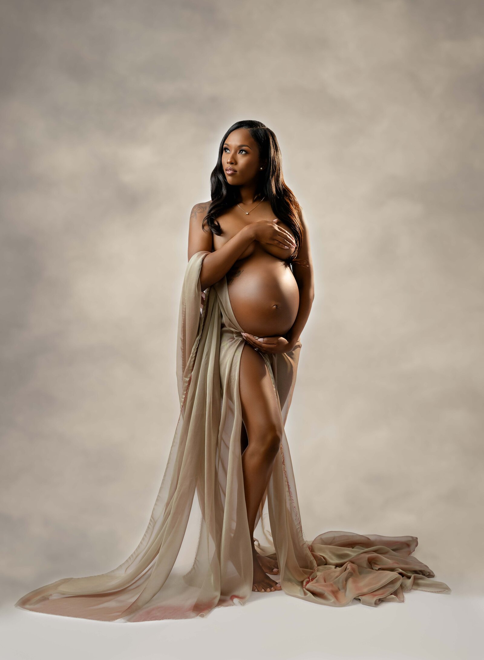 Pregnant women holding chest and belly and wrapped in gold drape, looking over shoulder. Photographed at Atlanta studio.
