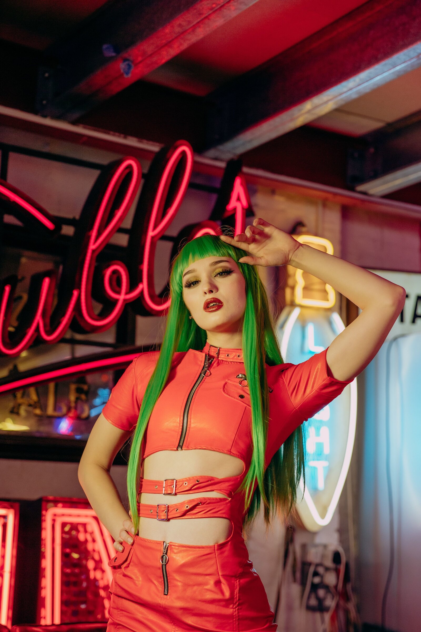 Saida wearing a red leather dress with thigh high boots and a neon green wig, infront of a neon sign saying "trouble"