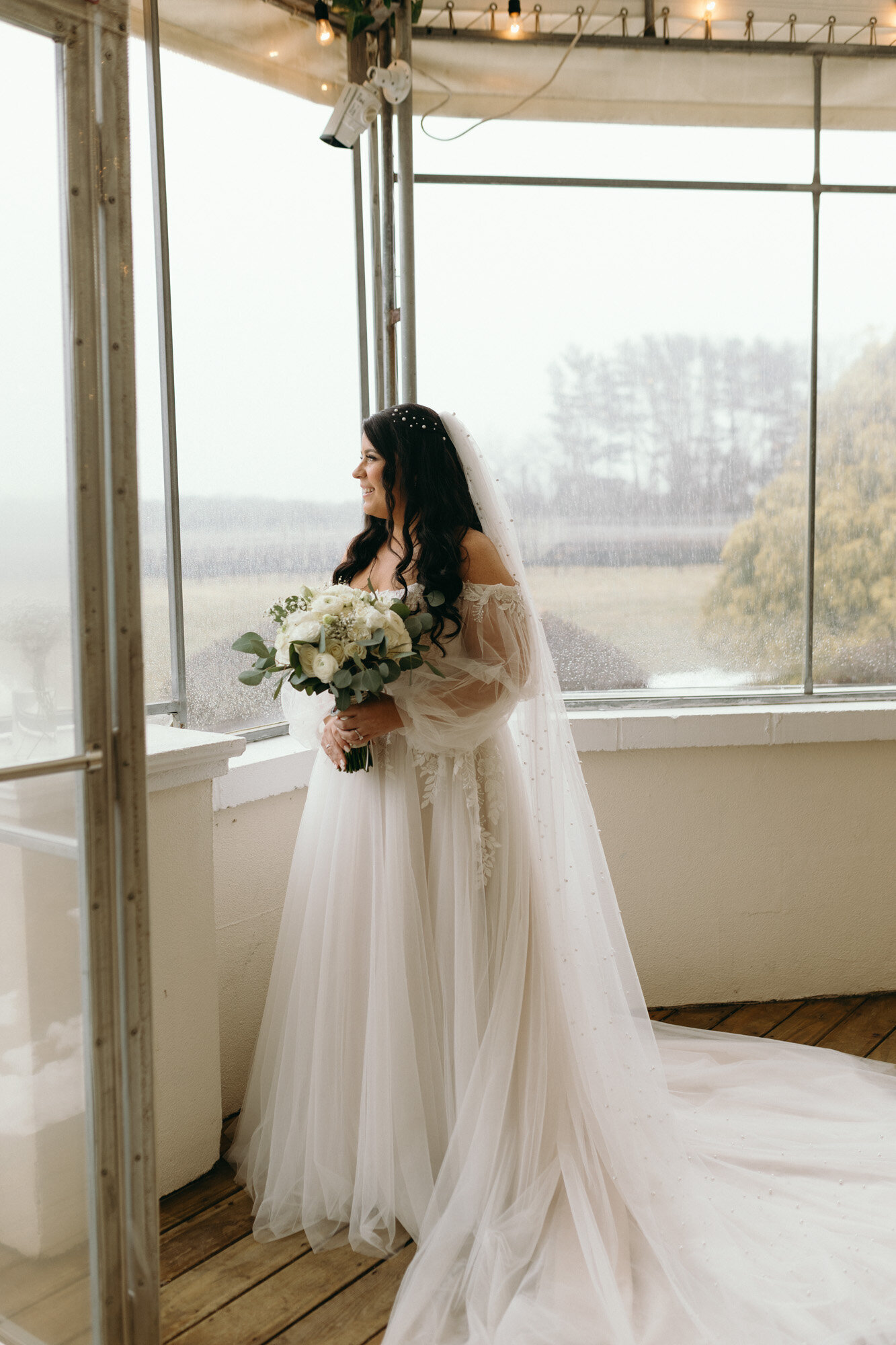 bride in a long, flowing white wedding dress and veil stands indoors in front of large windows, holding a bouquet of white flowers