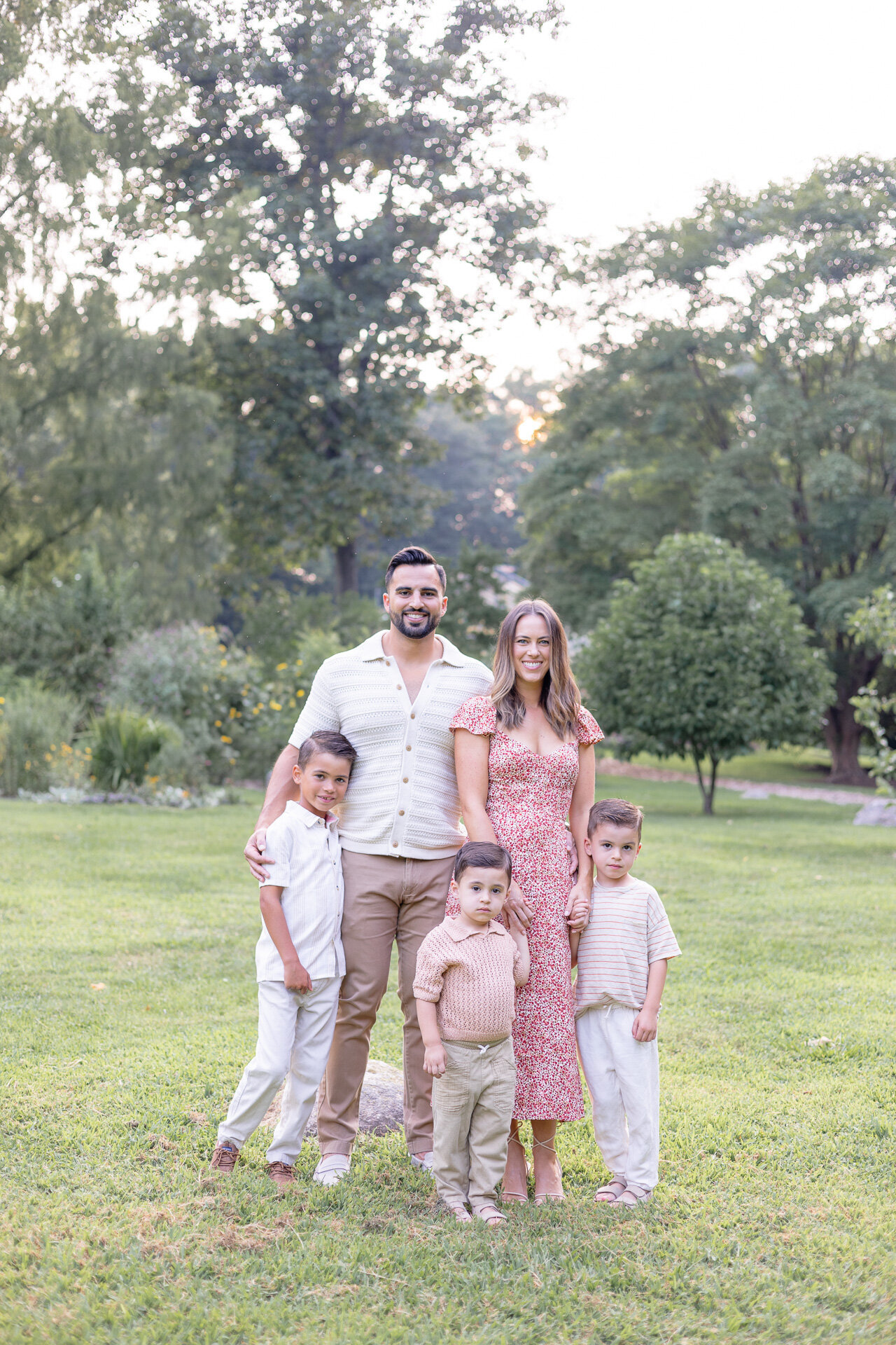 Family of five posing together in a sunlit field at golden hour during an outdoor family photography session in Fairfield County, CT. Natural and heartfelt portrait showcasing parents with their three young sons in a lush summer setting.