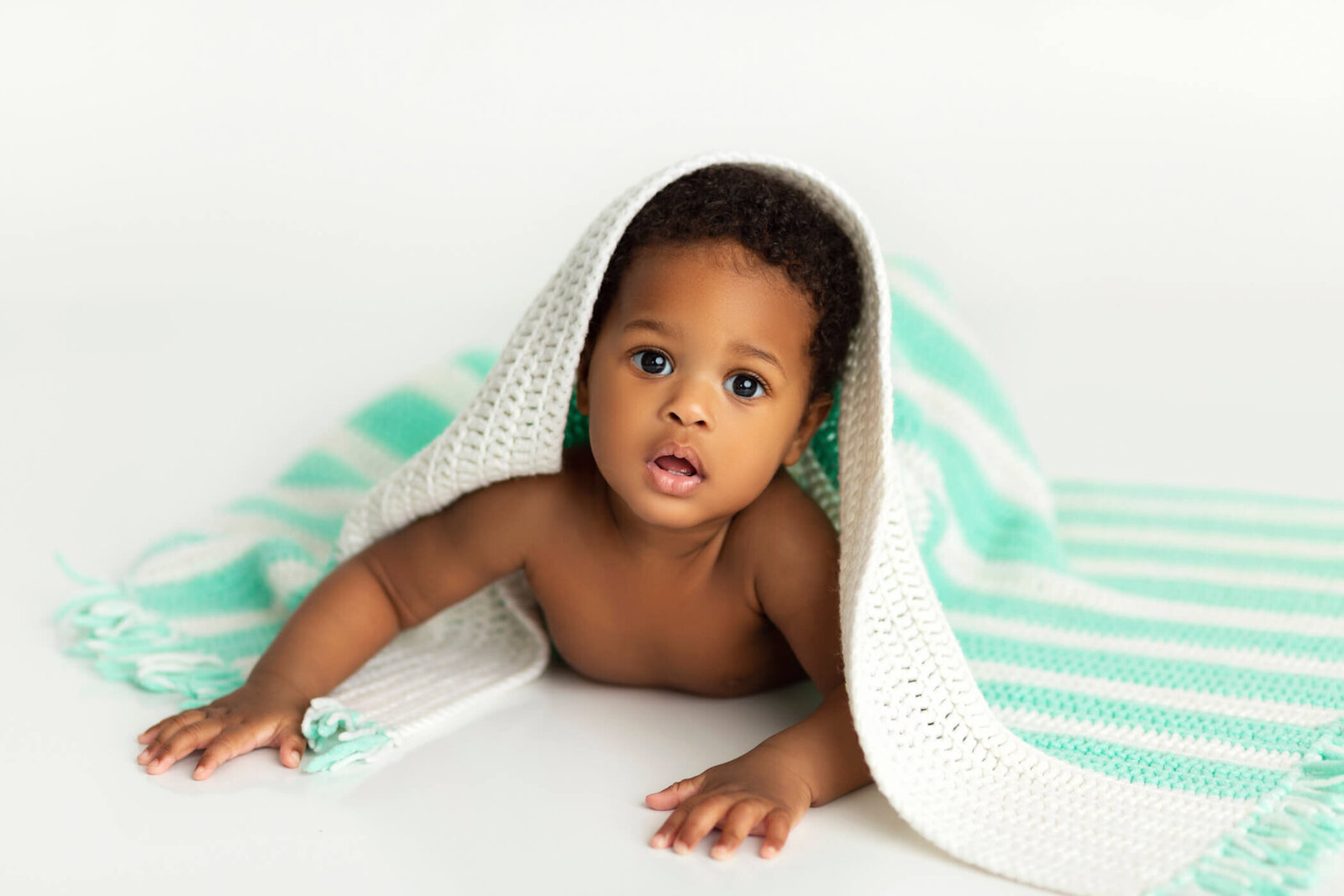 A cute baby with short, curly hair and big, expressive eyes lies on their stomach on a white surface. The baby is partially covered by a soft, white and mint green striped knit blanket, which drapes over their back and head like a cozy tent. The baby looks curiously at the camera with an open mouth, creating an engaging and heartwarming scene. The background is a clean, bright white, emphasizing the baby's smooth, dark skin and the soft textures of the blanket.