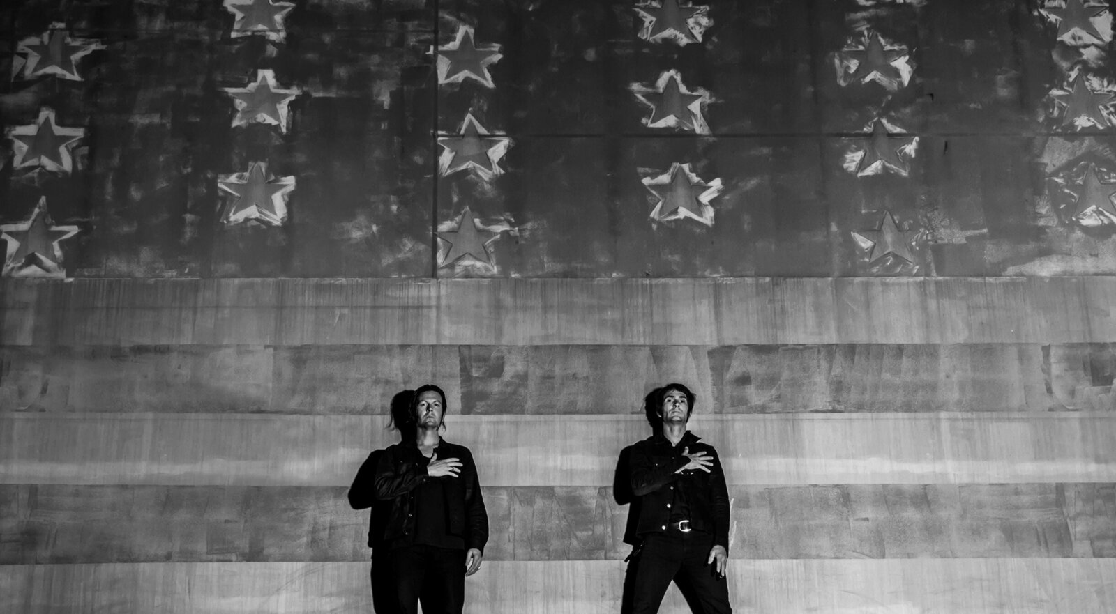 Band portrait Los Angeles Walking Papers both members standing in long shot against American Flag painted on wall hands across their hearts black and white