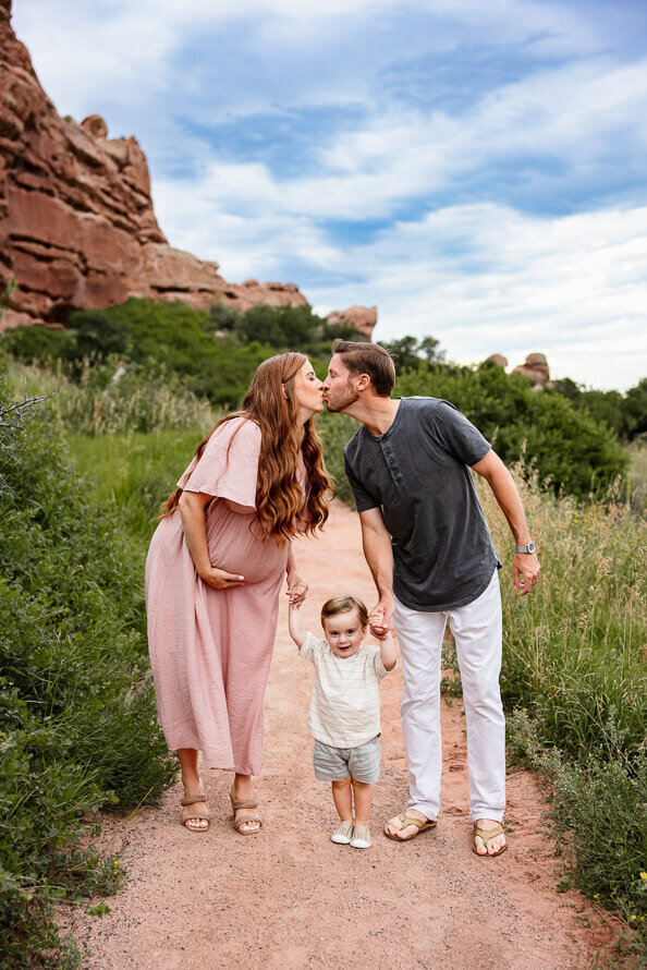 Pregnant mom kisses her husband while holding hands with their toddler at South Valley Park in Colorado.