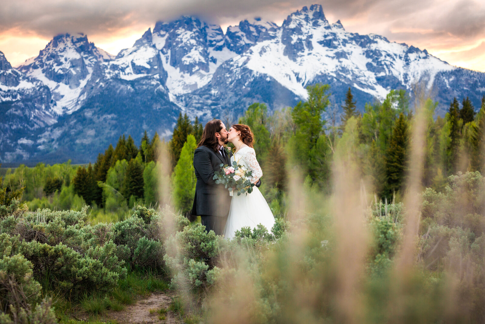 Bride and groom kiss behind sagebrush at their jackson hole wedding at schwbachers landing. The mountains are deep blue and the sky is peach and orange. Perfect summer sunset wedding in Grand Teton National Park.