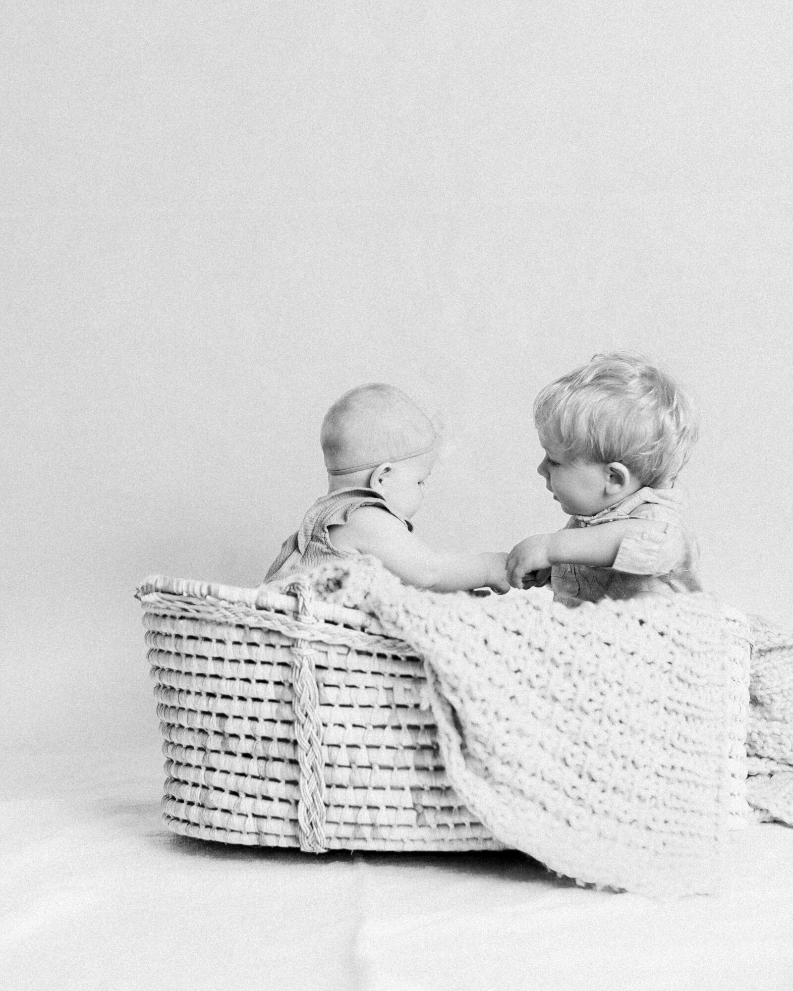 Toddler and baby siblings sitting in a moses basket together playing and holding hands by NH newborn photographer Fieldstone Studio.