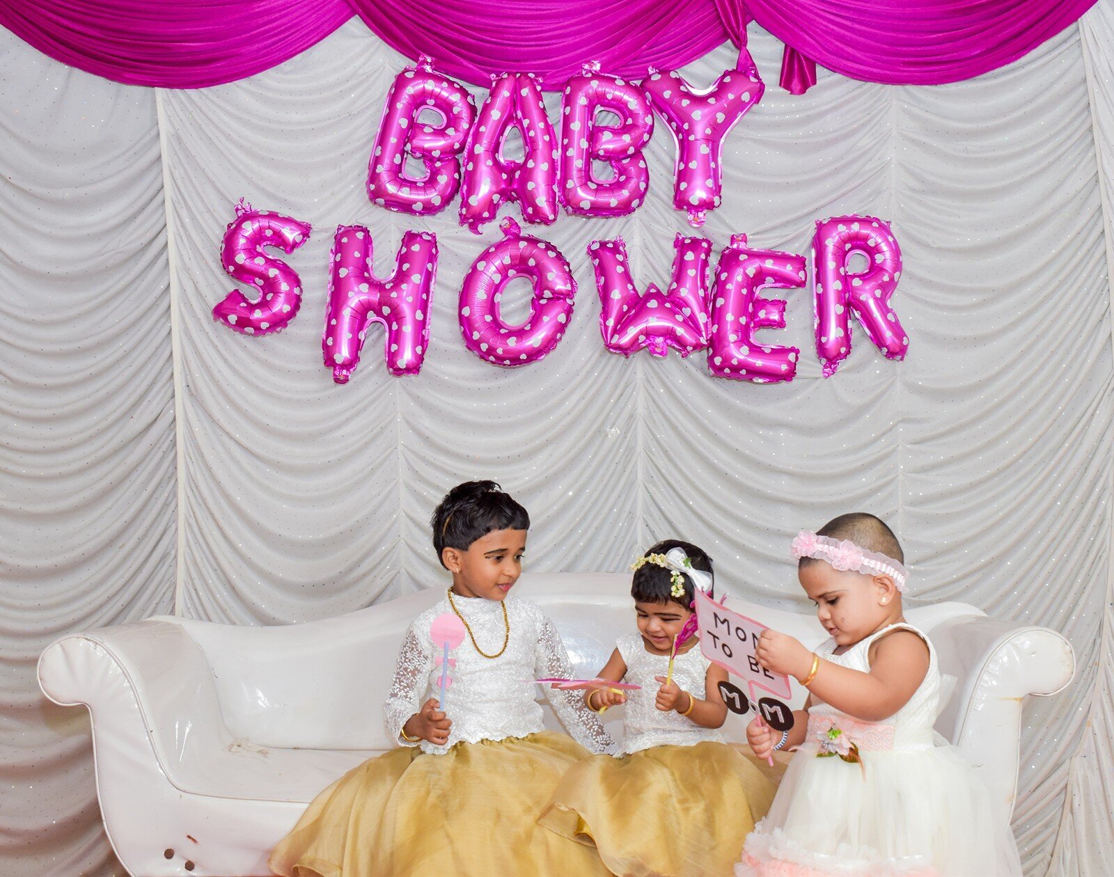 Kids at a baby shower sitting under pink “BABY SHOWER” balloon backdrop