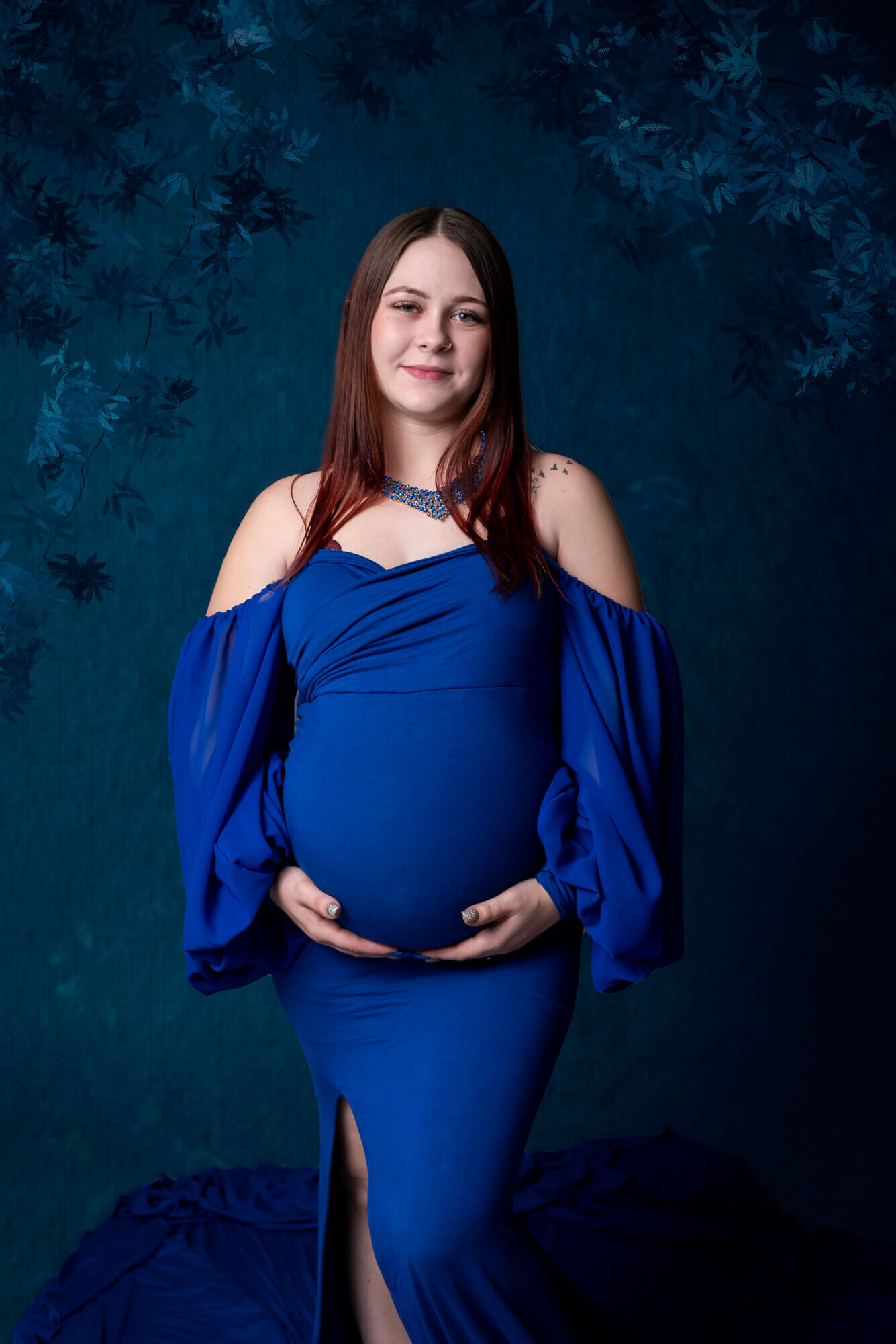  Radiant mother in a royal blue off-the-shoulder gown stands in front of a rich blue backdrop, her hands gently resting on her belly.