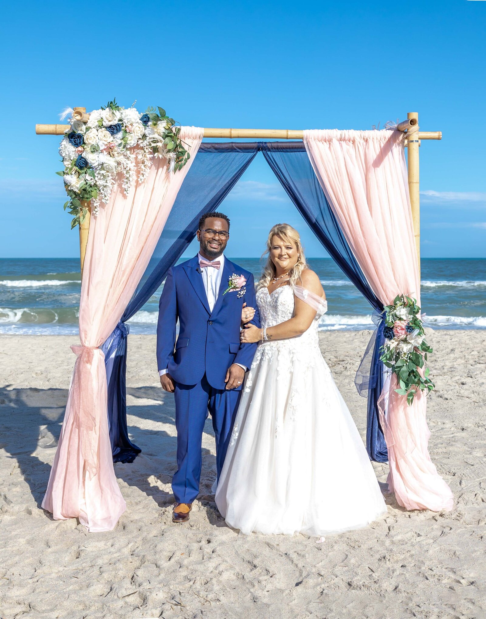 The bride and groom pose under a bamboo ceremony arbor draped with pink and navy fabric, adorned with flowers, captured during a Carolina Beach, NC wedding, blending coastal charm and vibrant elegance.