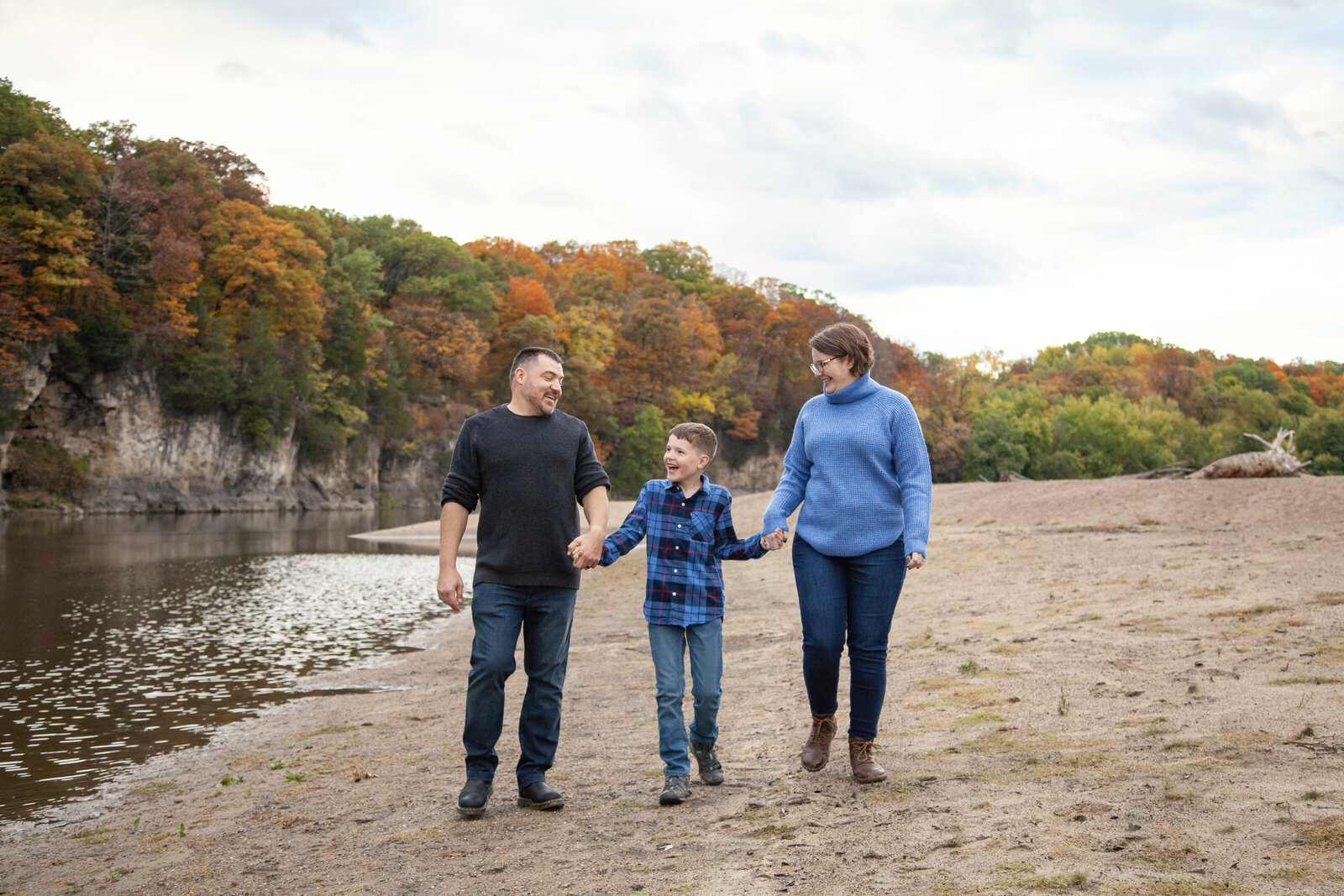 Son holding each parents hand as they walk in sand next to river, fall trees in background.
