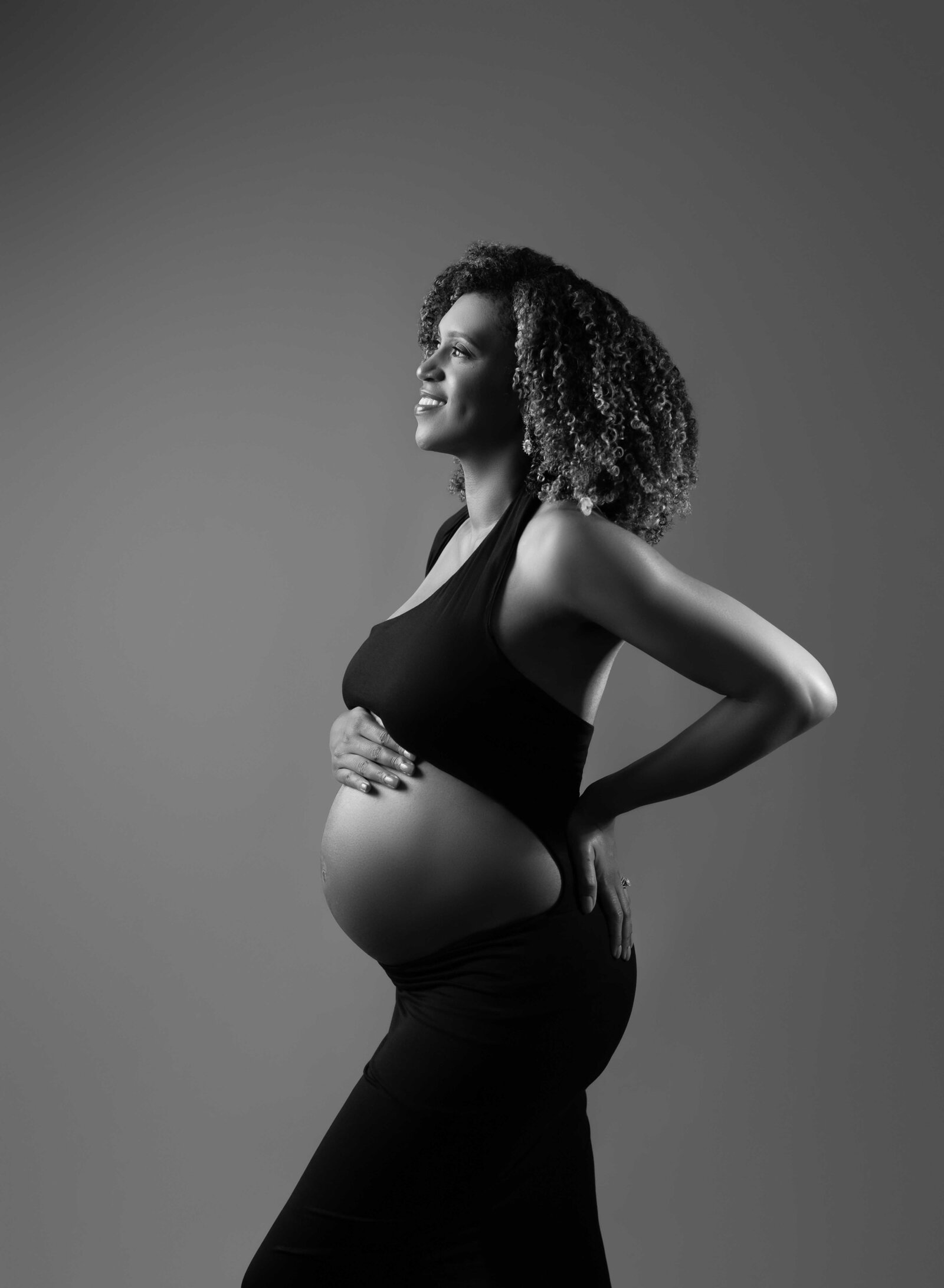 Black women in black dress smiling with hand on baby bump. Photo taken by Jenni Nikki Atlanta Maternity Photographer.