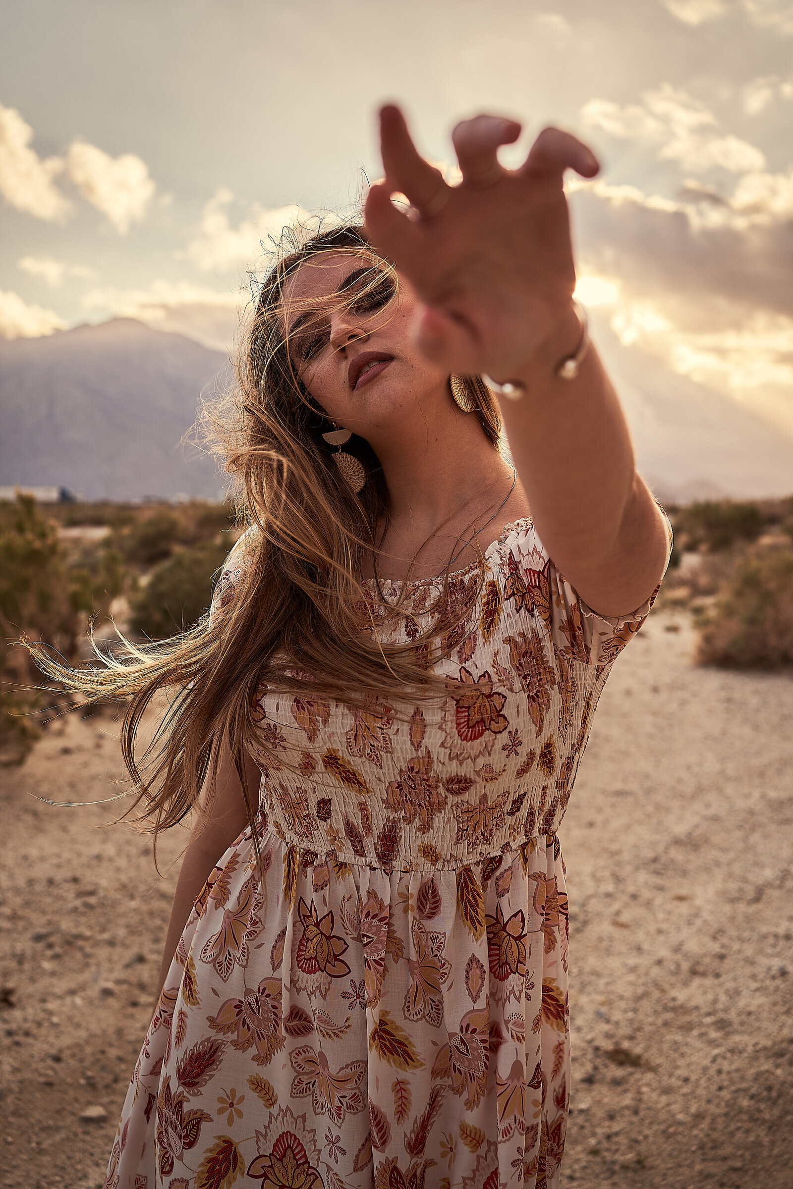 woman stands in the middle of portrait with her left arm reaching out towards the camera wearing a floral long dress and big gold feather earrings styled by free people