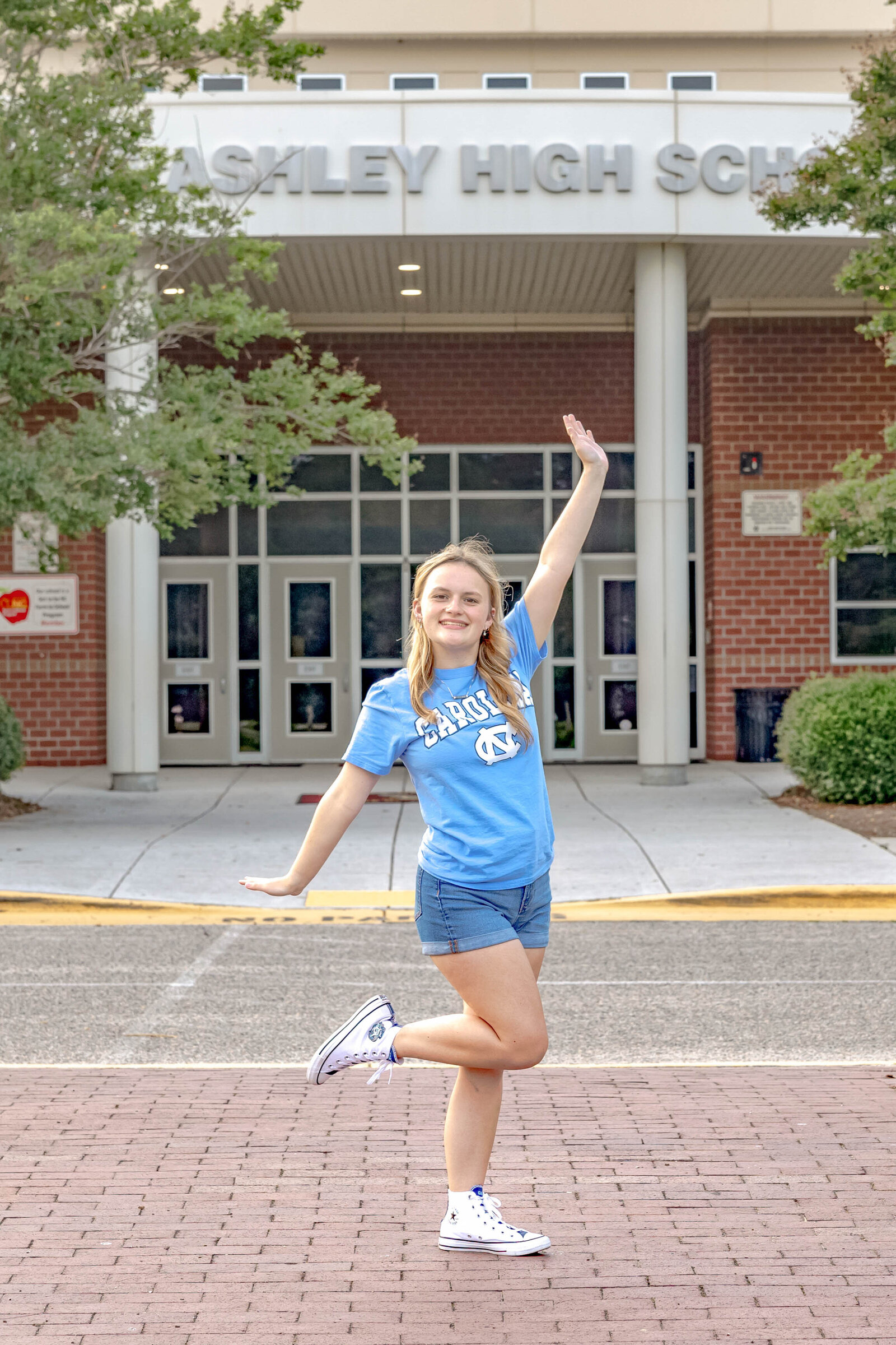 A high school senior in a Carolina shirt and shorts posing playfully in front of Ashley High School in Wilmington, North Carolina. This energetic photo highlights the excitement of senior year, ideal for vibrant and fun senior photography.