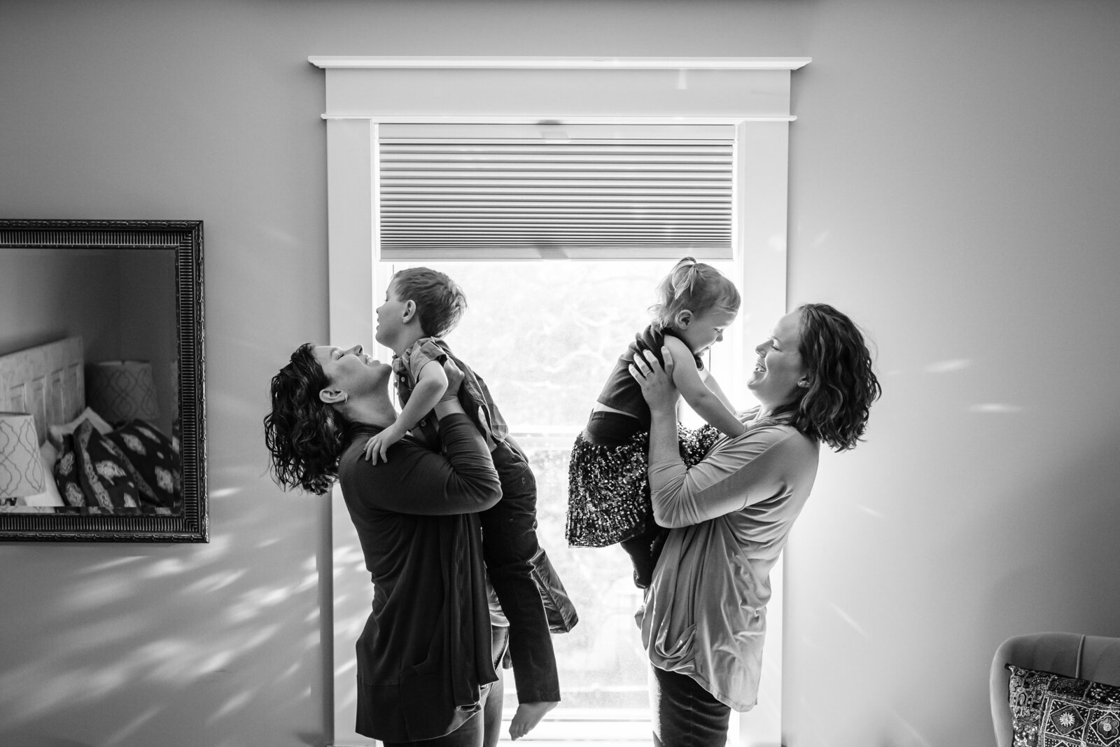 A black-and-white photo of two mothers holding their young children in an embrace, standing in a softly lit room.