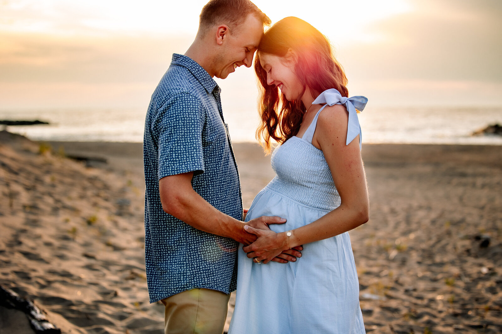 Couple stands with foreheads together looking down at mother's belly against a golden sunset