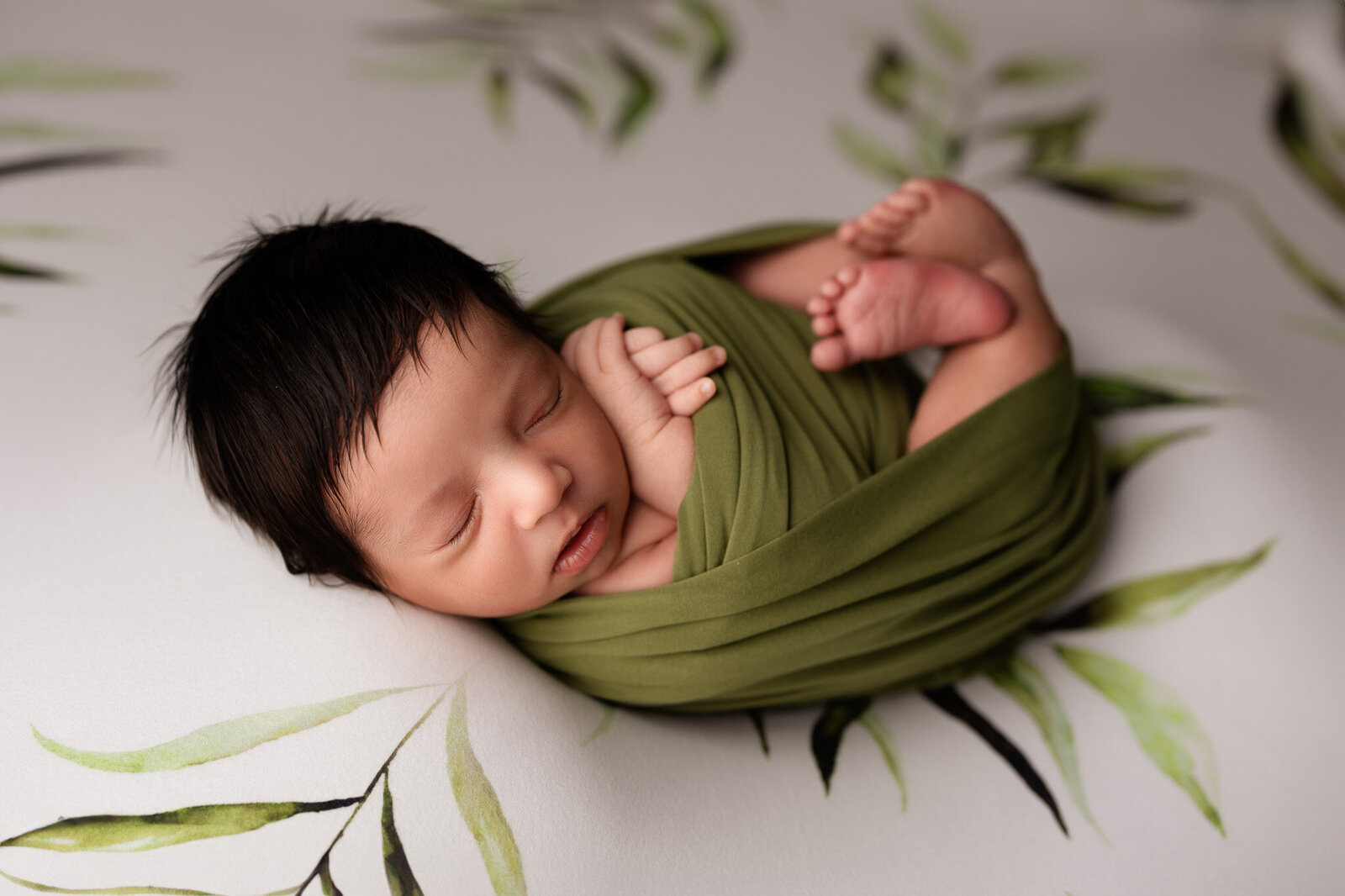 Baby boy with a full head of black hair, wrapped up in an olive green wrap and posed on a backdrop with a green leaf pattern