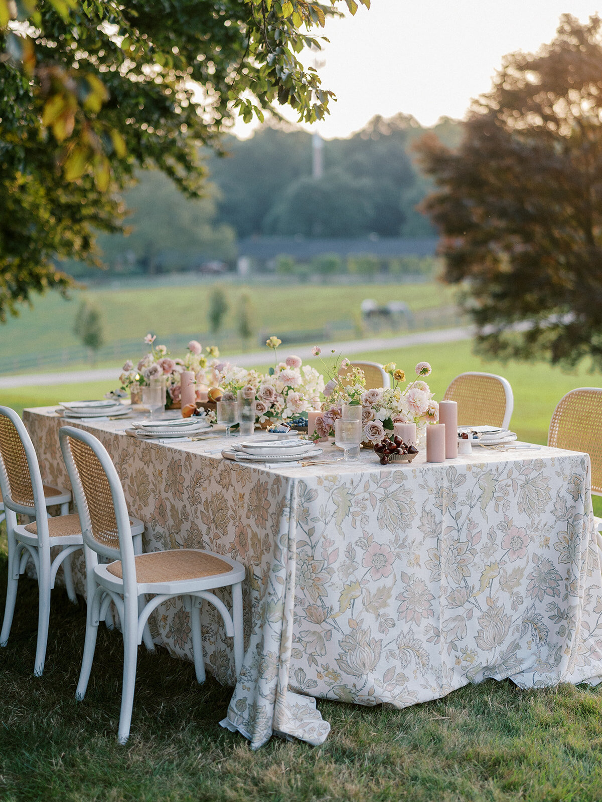Long table with floral-patterned linen with blush pillar candles, fresh fruit and blush, taupe, cream and mauve wispy floral compotes going down the length of the table.