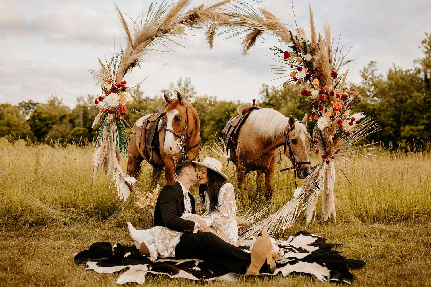 Bride and groom seated on a blanket in a field, surrounded by horses and a floral arch